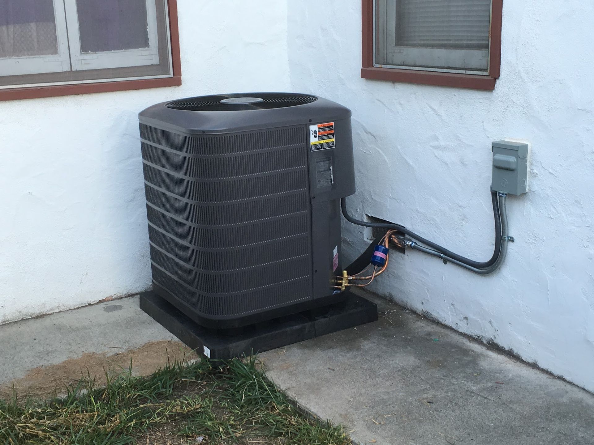 Air conditioning unit outside a building, black unit against white wall, electrical box visible.