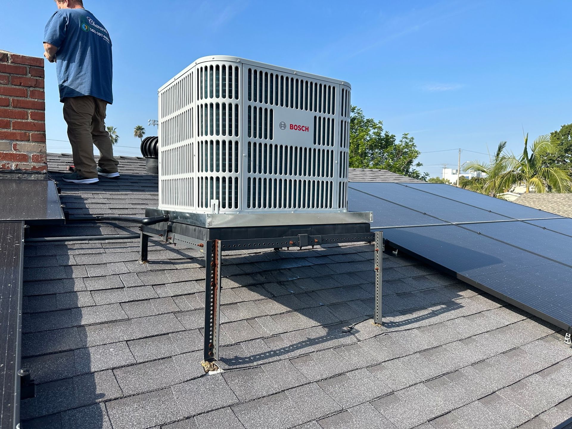 Air conditioning unit on a roof with a technician standing near a brick chimney and solar panels.
