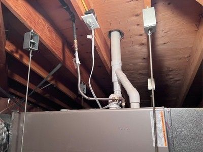 Furnace in an attic with white pipes, electrical wiring, and wooden beams.