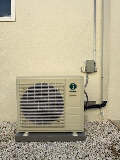 Outdoor air conditioning unit against a beige building wall, with electrical conduit and a gravel base.