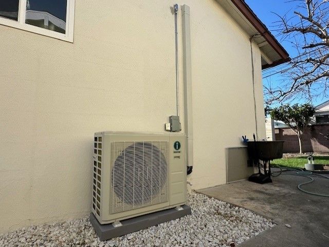 Outdoor air conditioning unit mounted on a concrete base against a beige wall, gravel below.