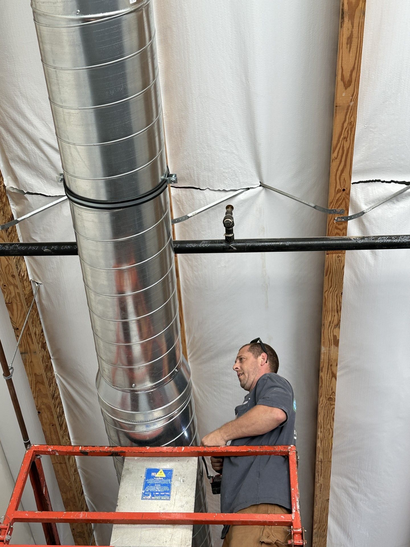 Man on lift installing ductwork in a building. The man wears a gray shirt and khaki pants.