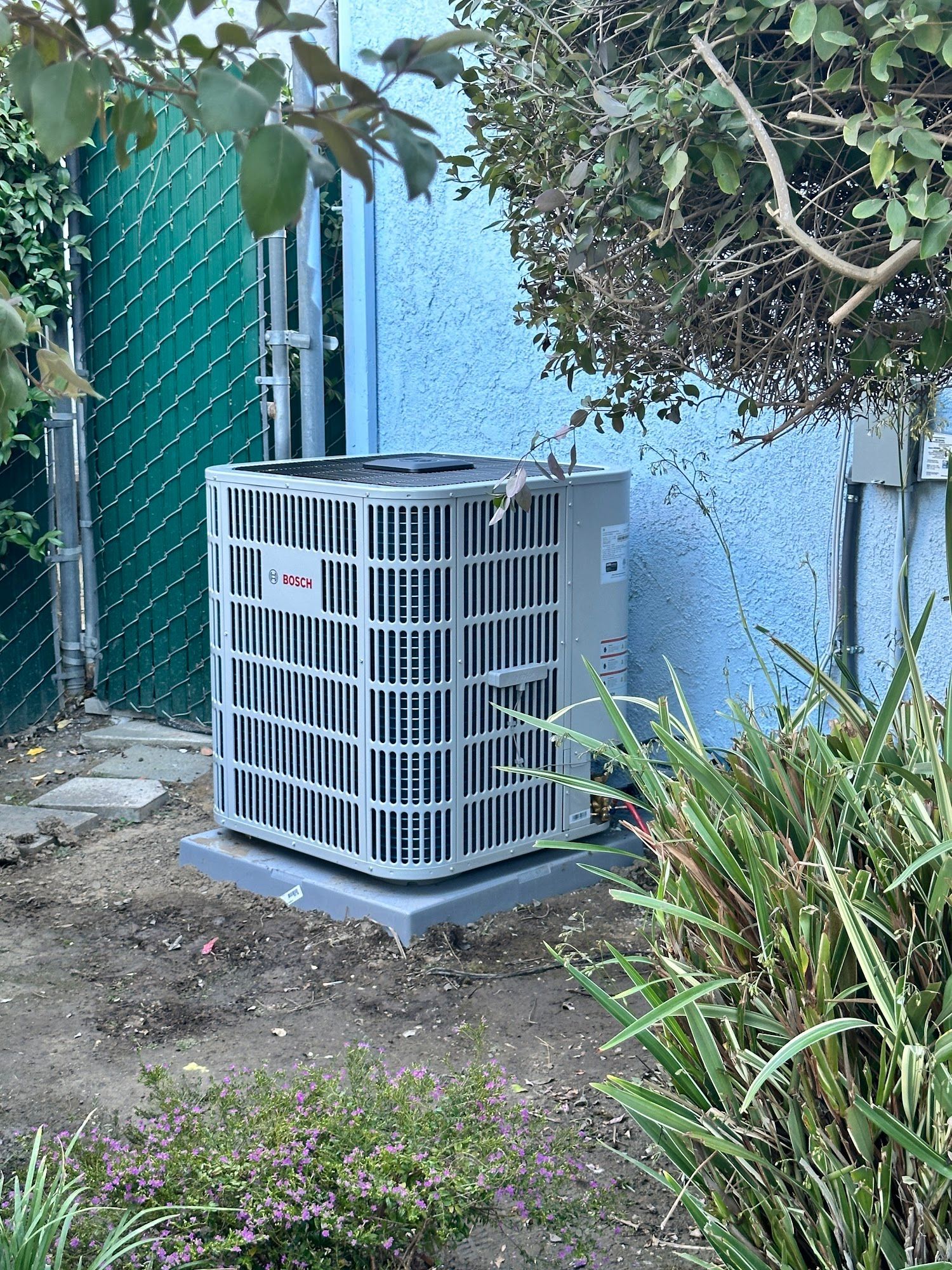 Air conditioning unit outdoors, next to blue wall and green foliage.