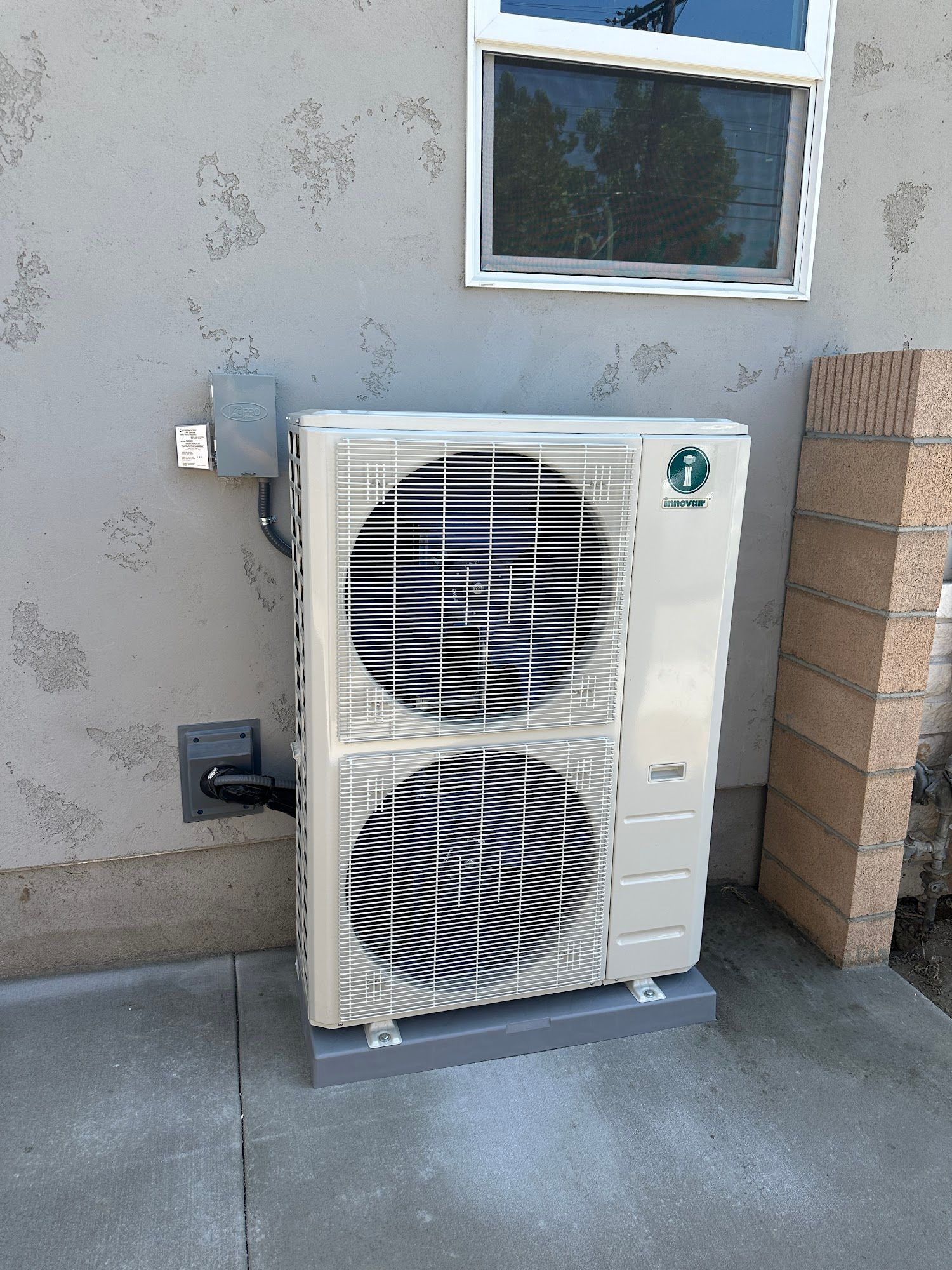 Air conditioning unit mounted on a gray base against a stucco wall near a window.