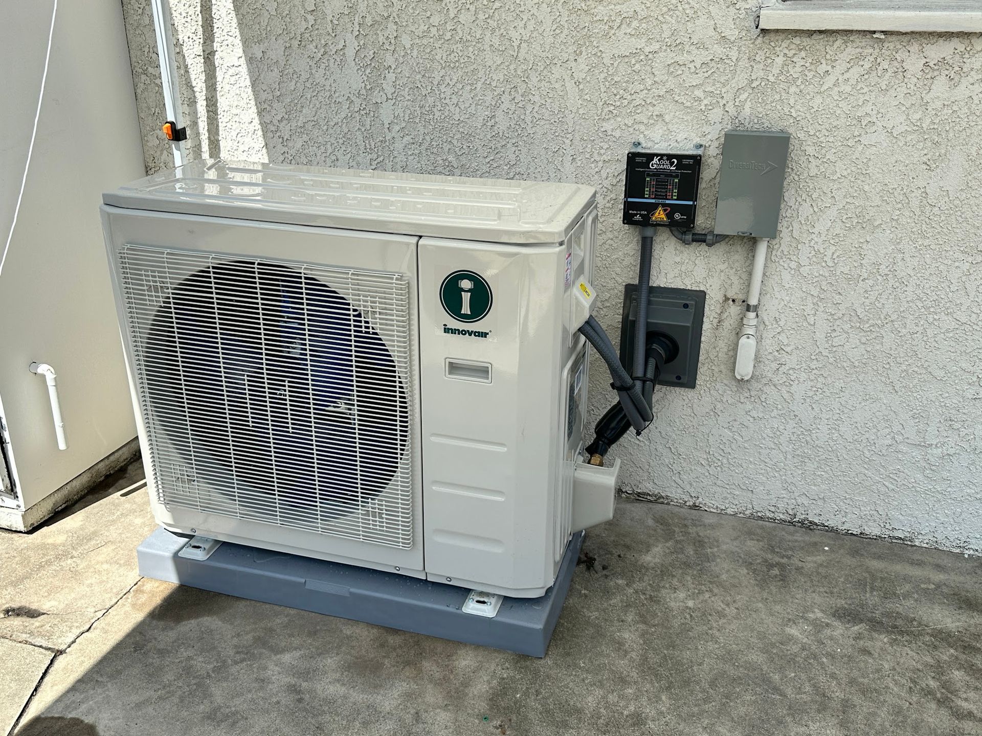Air conditioner unit mounted on a gray base, next to electrical components on a stucco wall.