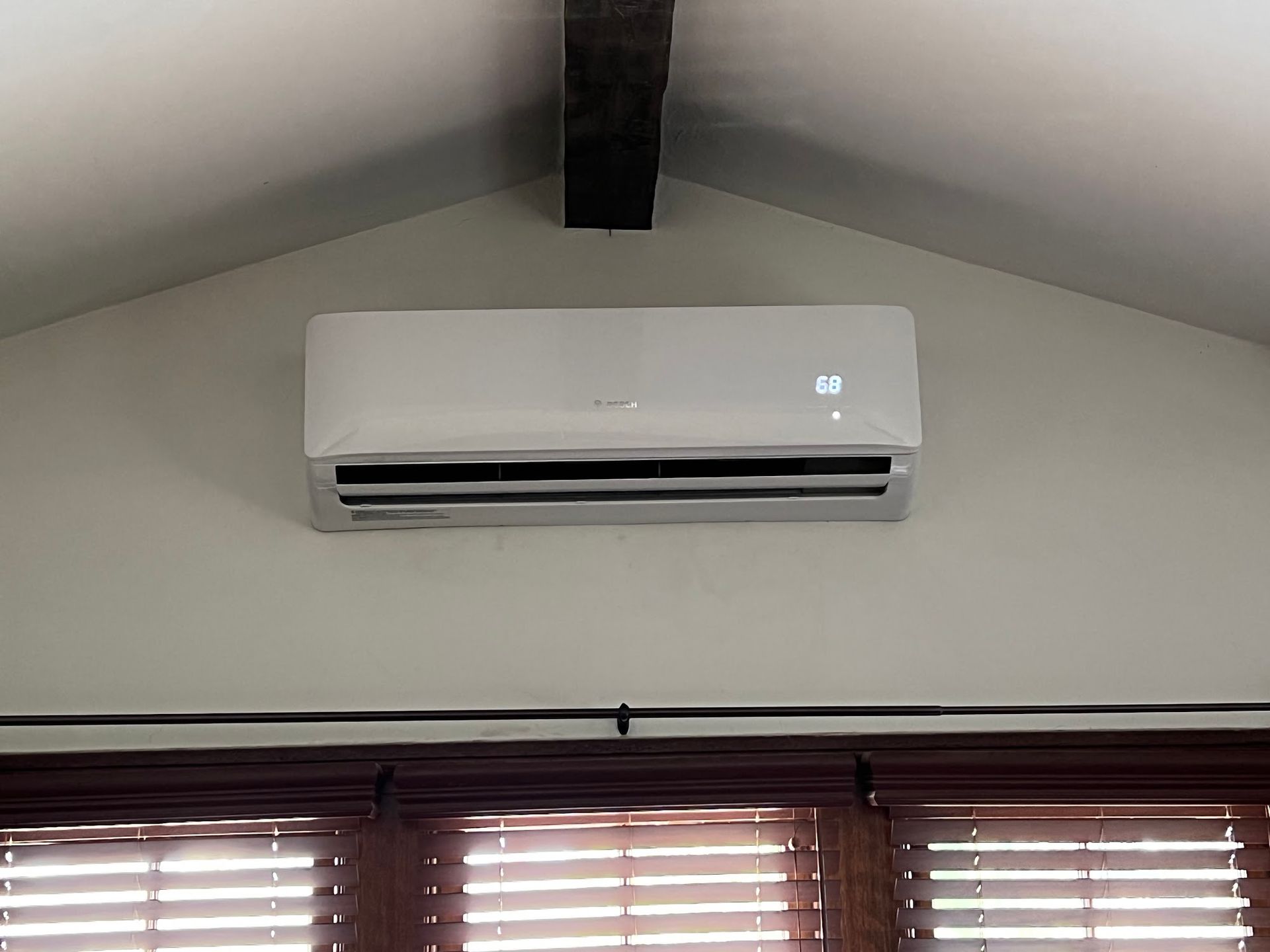 White wall-mounted air conditioner above dark blinds; a black beam is above it.