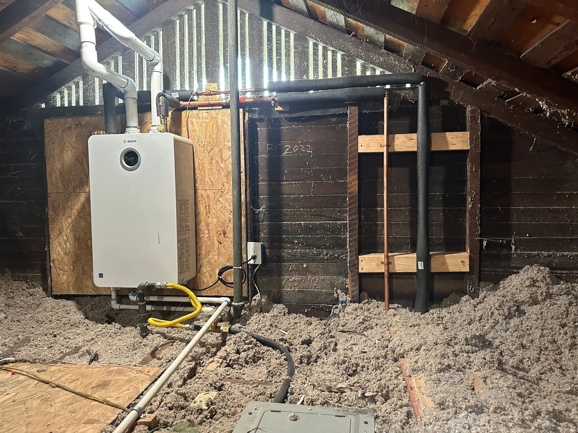 A small white water heater installed in an attic surrounded by insulation and wooden framing.