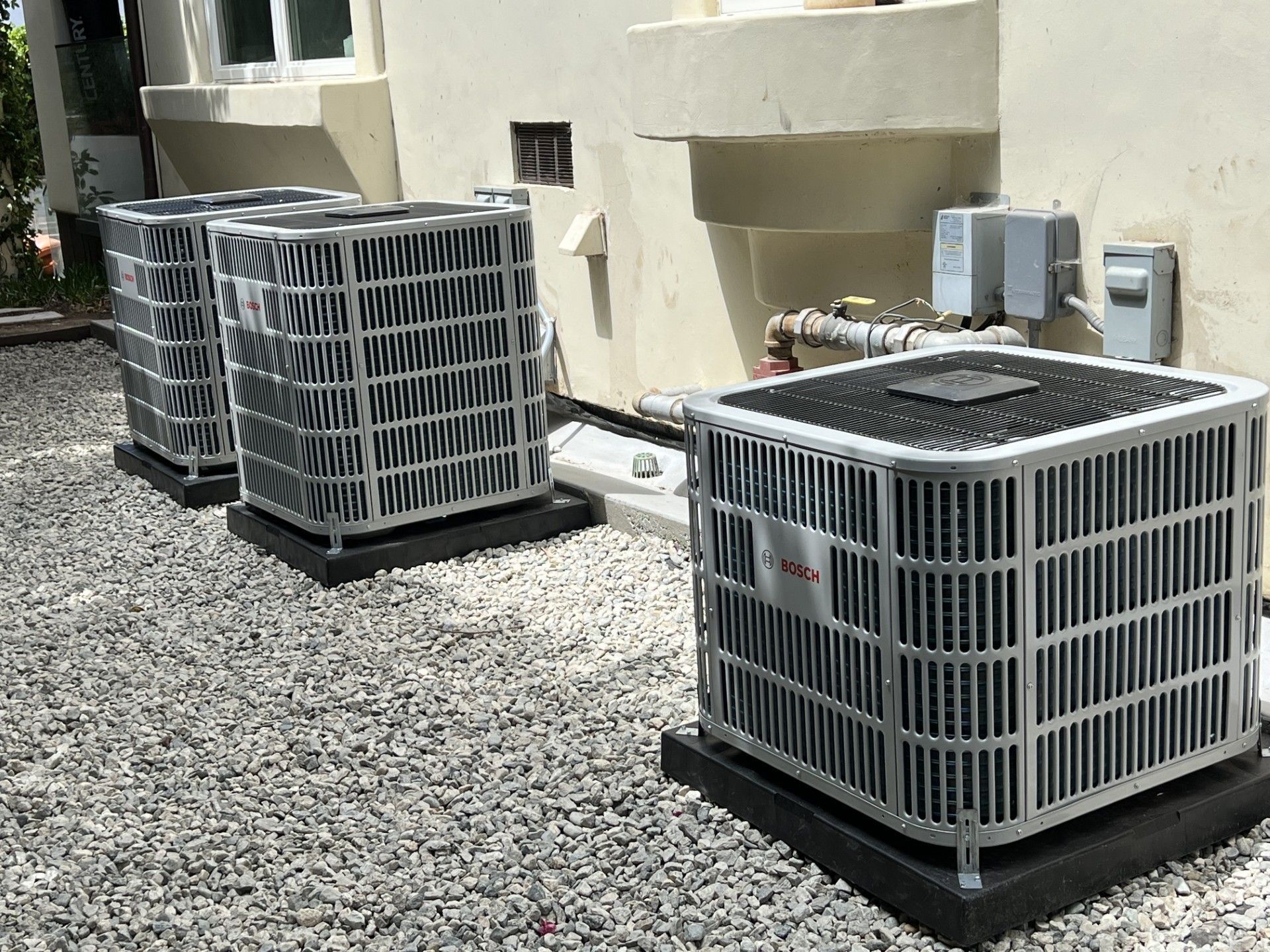 Three air conditioning units on gravel next to a building wall.