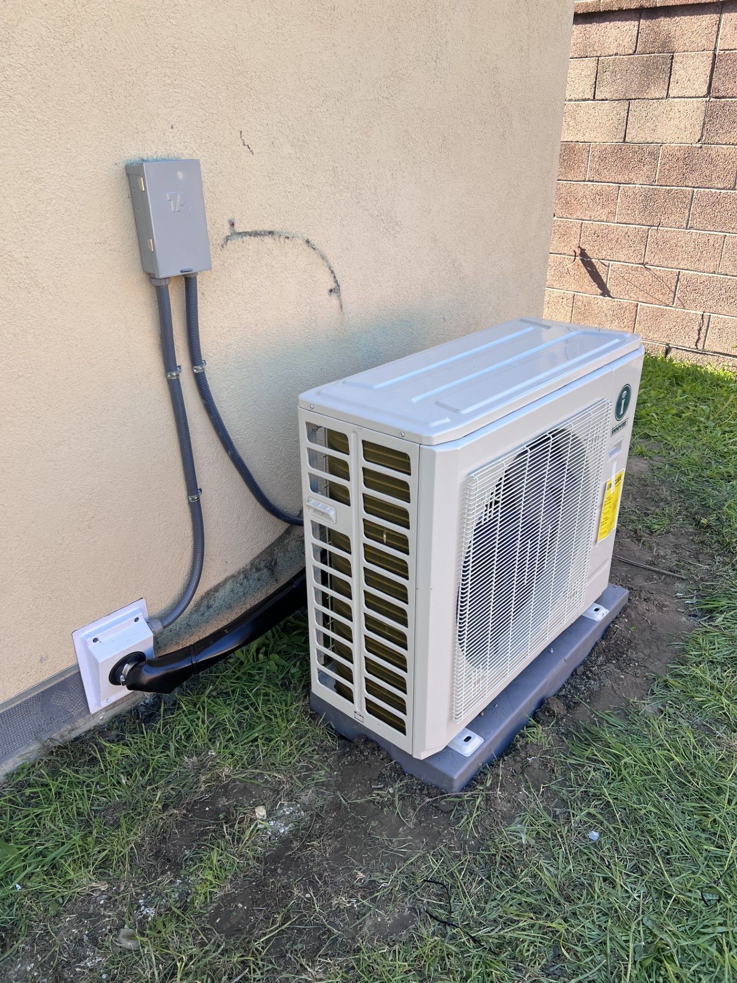 Outdoor air conditioning unit with electrical conduit on a beige stucco wall and green grass.
