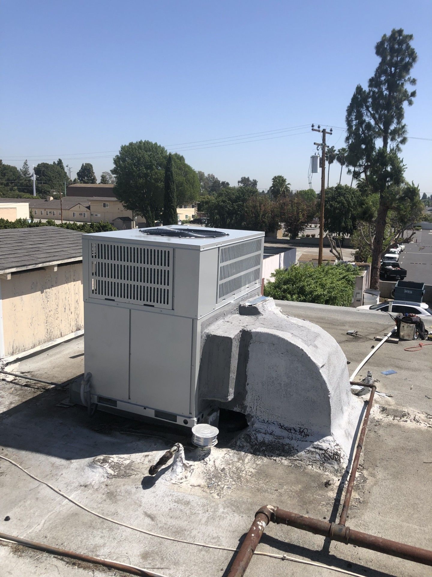 HVAC unit on a flat rooftop on a sunny day. Gray unit, white/gray roof, trees and buildings in the background.