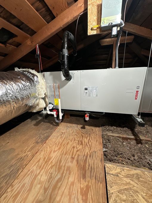HVAC unit in an attic with ductwork, insulation, and wooden planks.