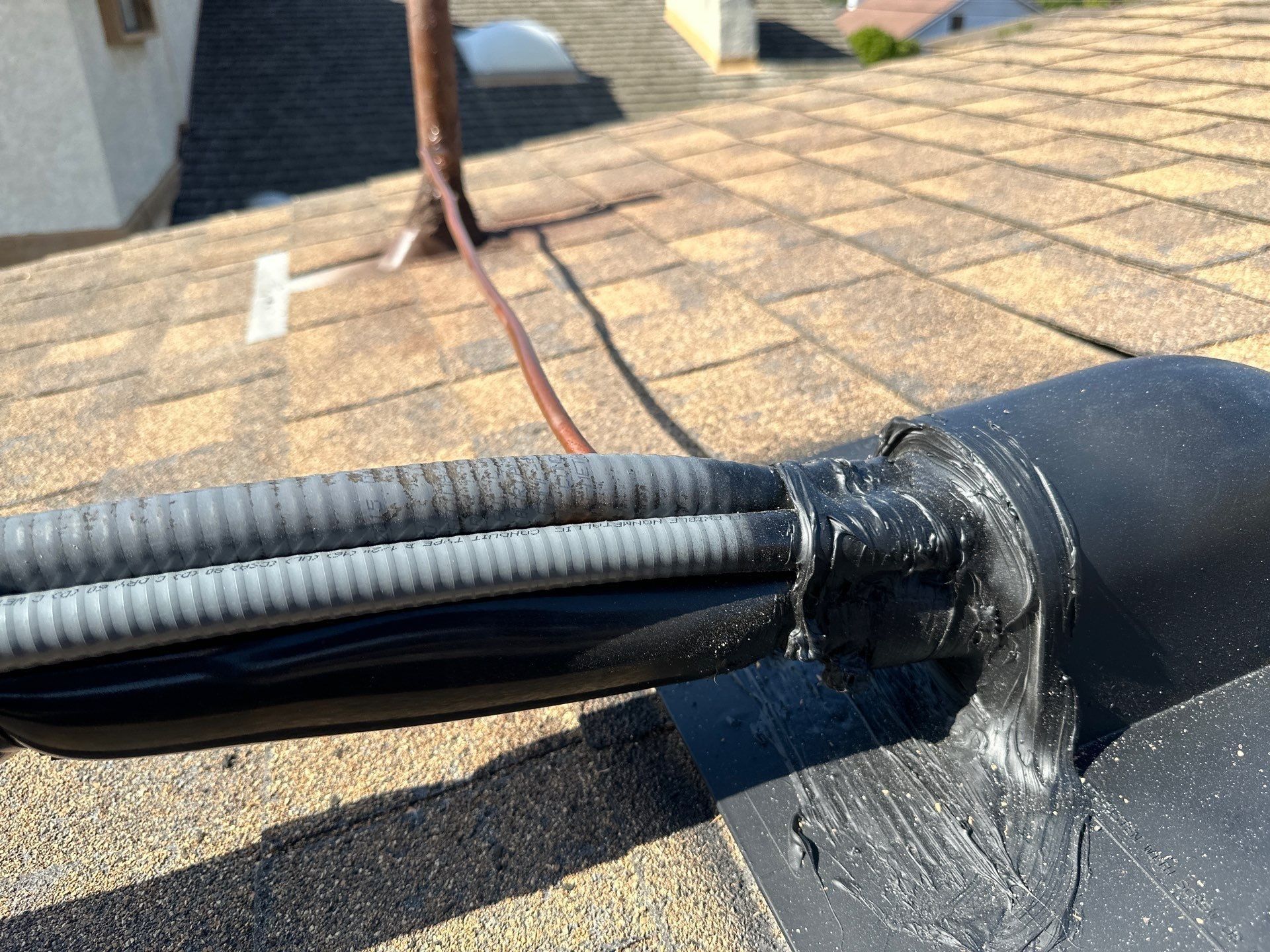 Close-up of a roof with shingles; wires sealed with black sealant around a vent pipe.