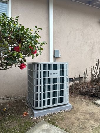 Central air conditioning unit on a concrete base next to a house with a bush and flowers.
