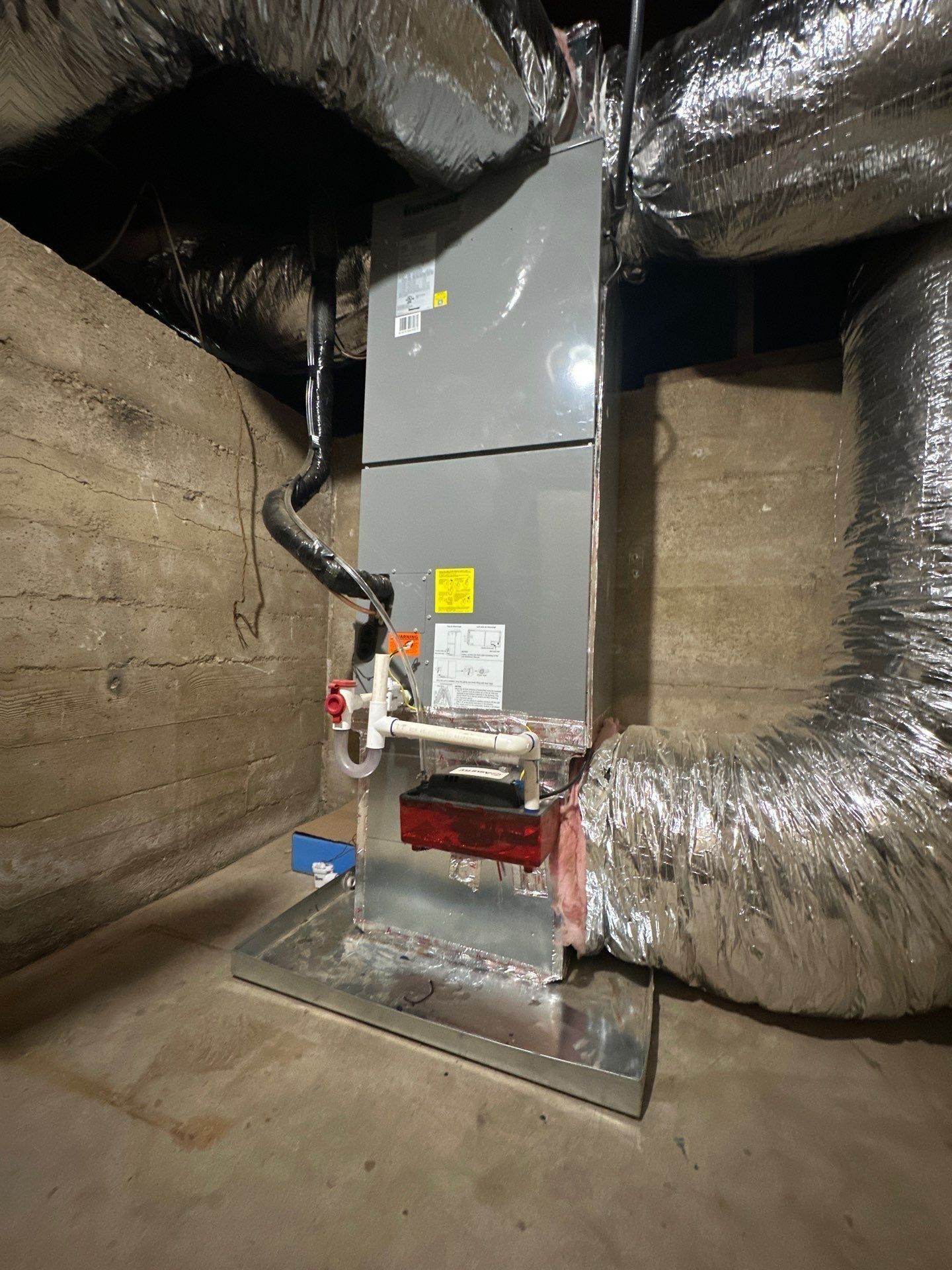 HVAC unit with ductwork in a basement setting. Grey metal box with piping, red components, and silver ducting.