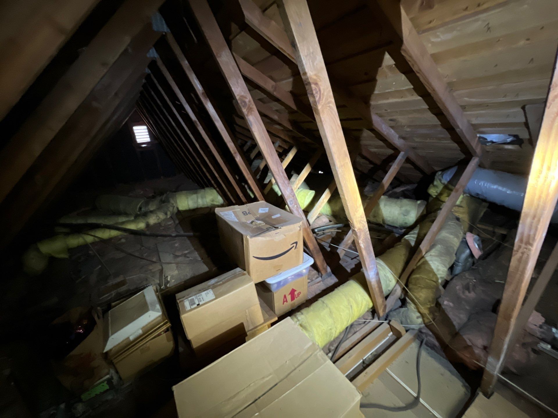 Attic interior with wooden rafters, insulation, and cardboard boxes. Dark, enclosed space.