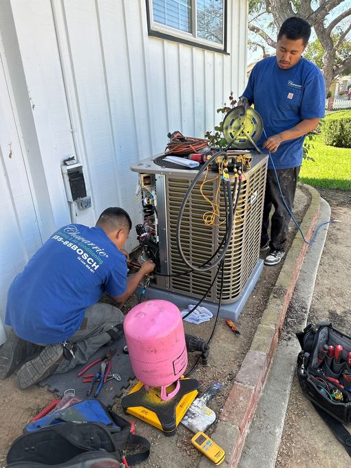 Two technicians in blue shirts repair an outdoor air conditioning unit next to a white building.