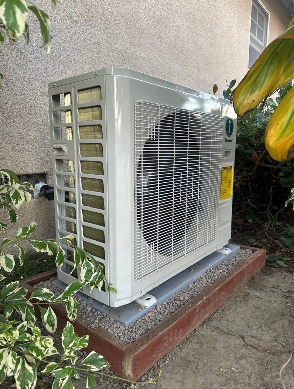 Outdoor air conditioning unit, light gray, sitting on a brick and gravel base next to a wall and foliage.