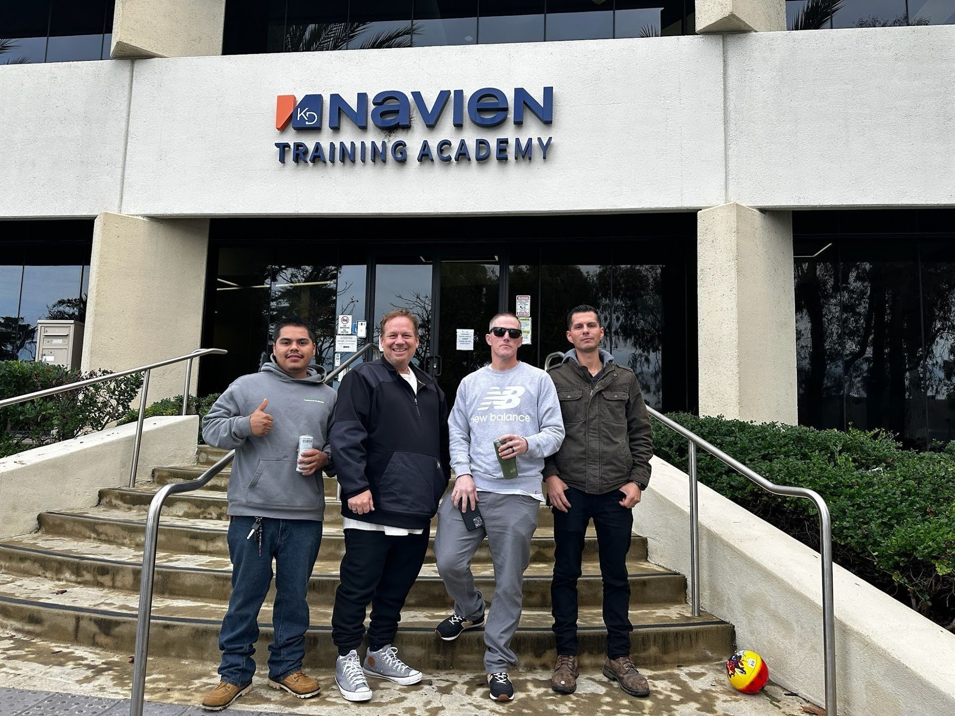 Four people stand in front of the Navien Training Academy building, posing on the steps, holding drinks.