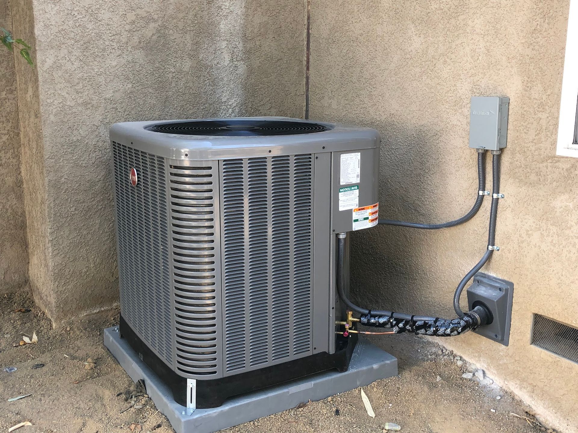 Gray air conditioning unit outside a stucco building, with electrical components and piping.