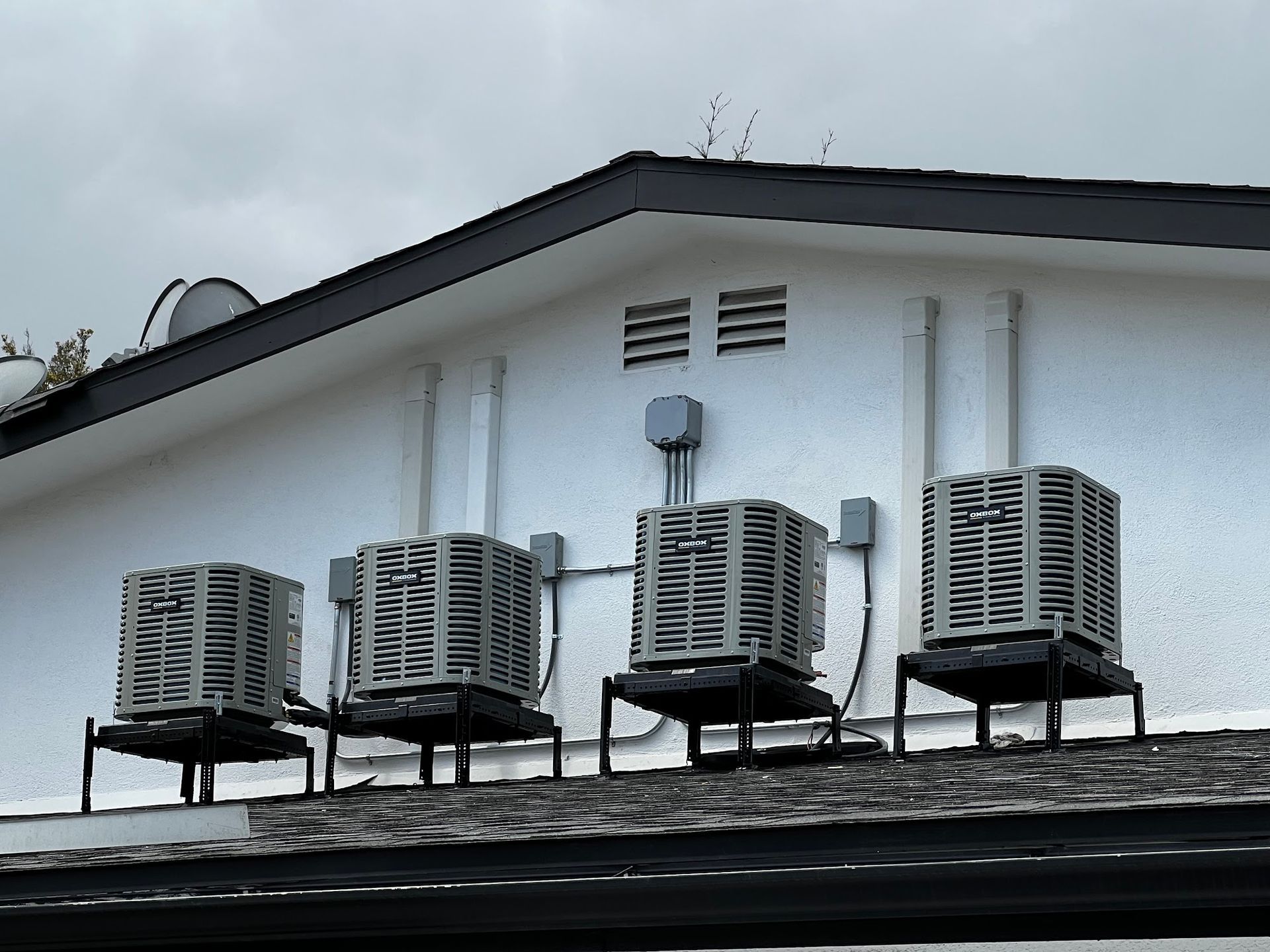 Four air conditioning units on a white building roof with vents and electrical boxes against a gray sky.