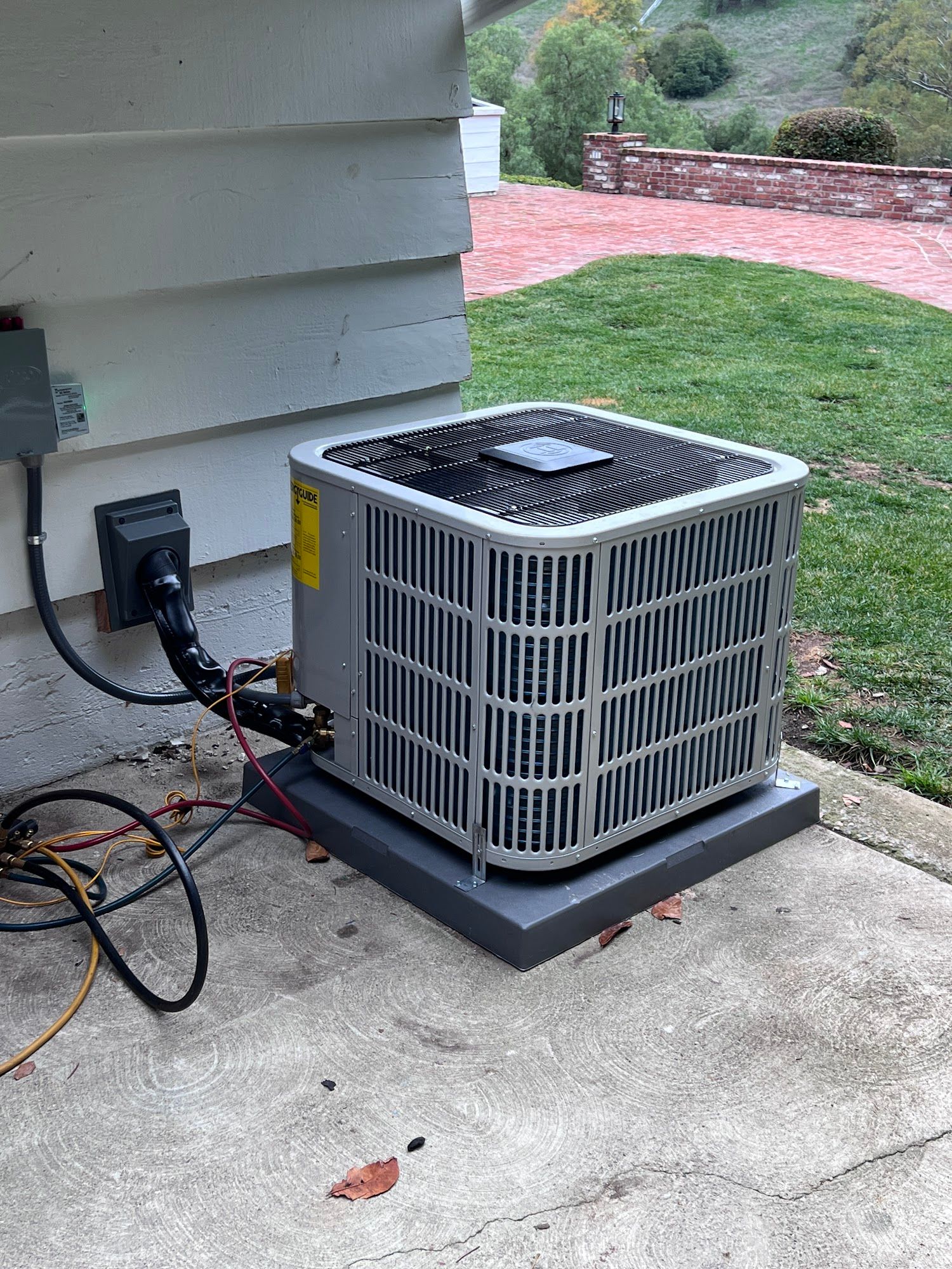 AC unit on a gray concrete pad next to a white house with electrical wiring; grass and a brick wall are in the background.