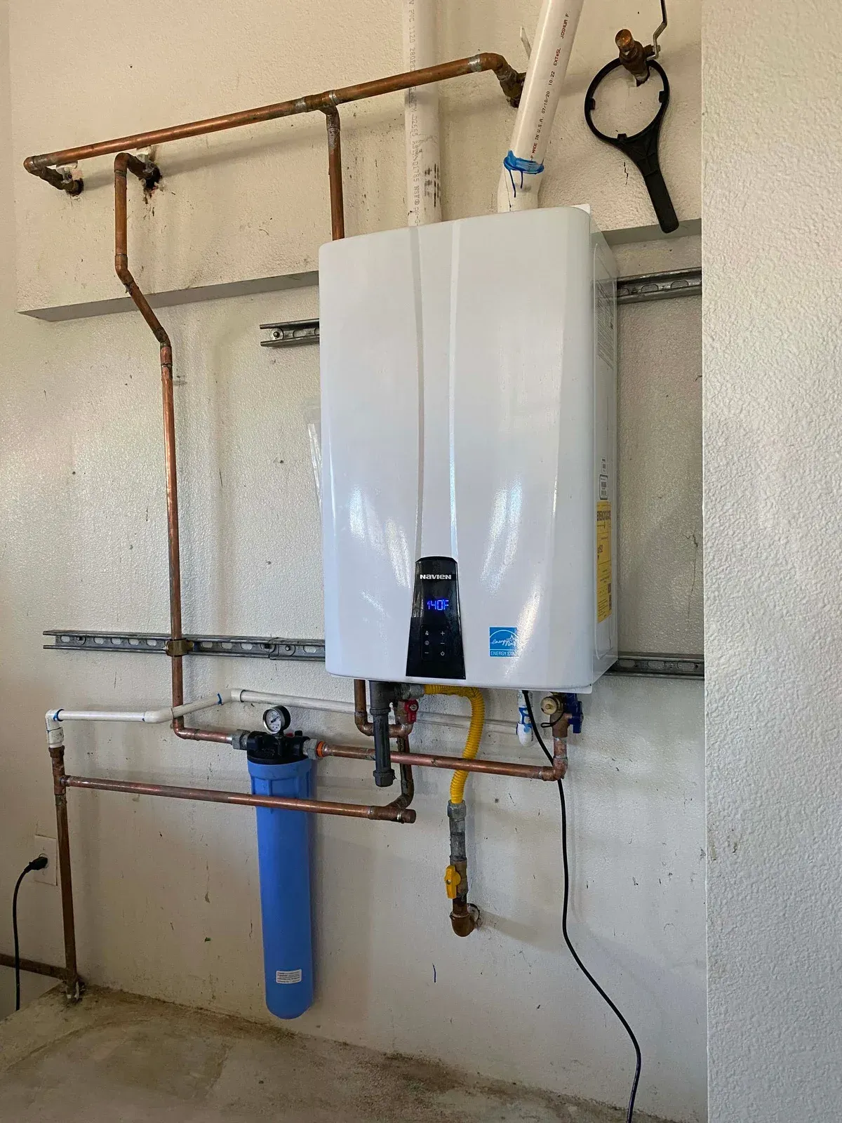 White tankless water heater mounted on a wall with copper pipes, a blue water filter, and a wrench.