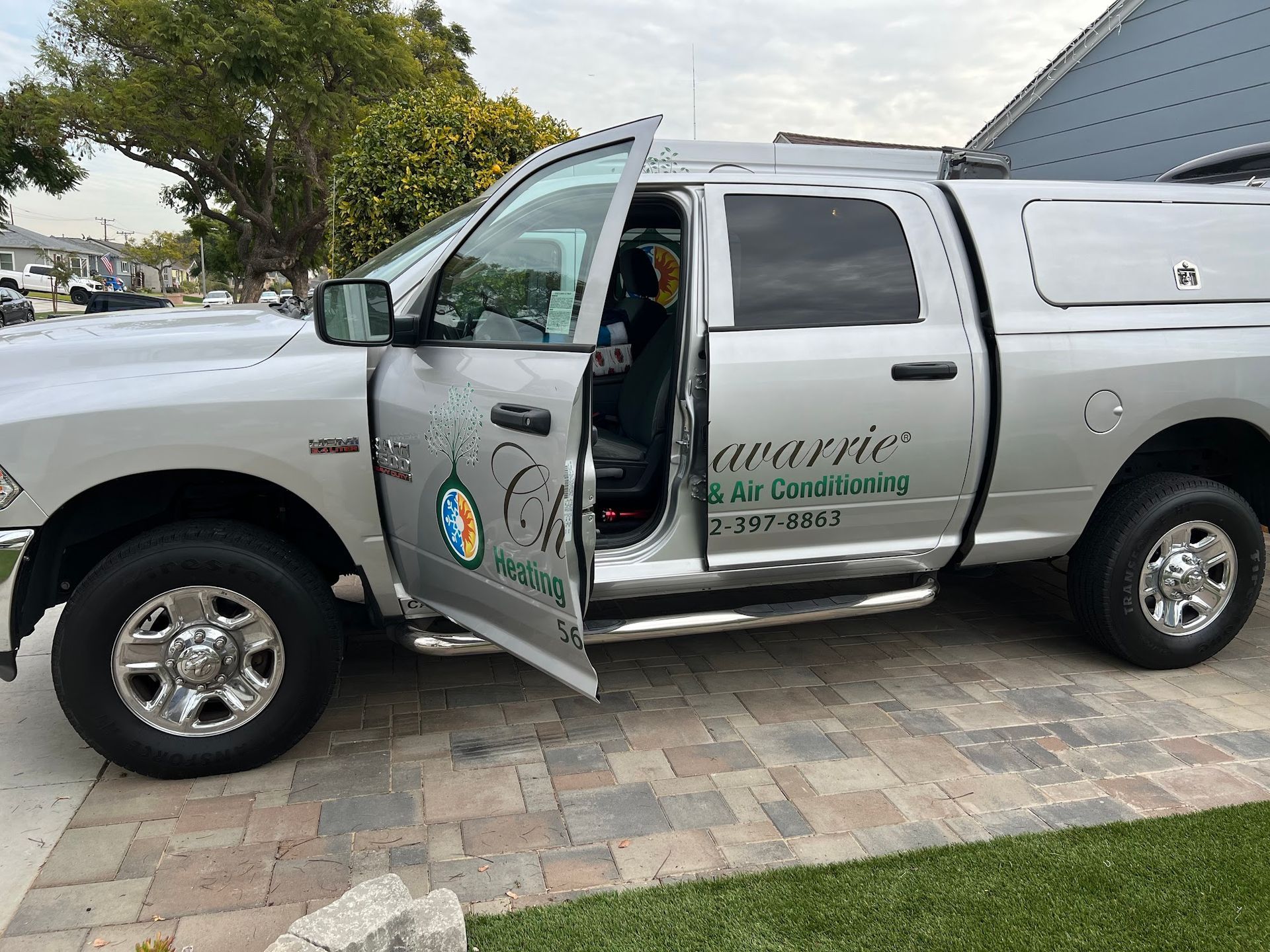 Silver pickup truck with company logo, parked on a brick driveway, open driver-side door.