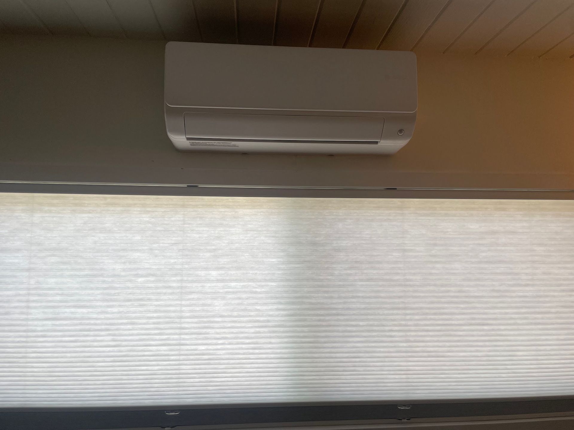 White mini-split air conditioner mounted above closed, light-colored pleated blinds.