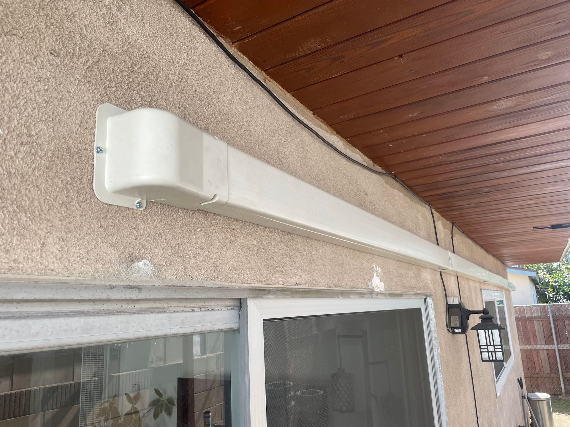 Beige awning mounted above a sliding glass door on a stucco building with a wooden overhang.