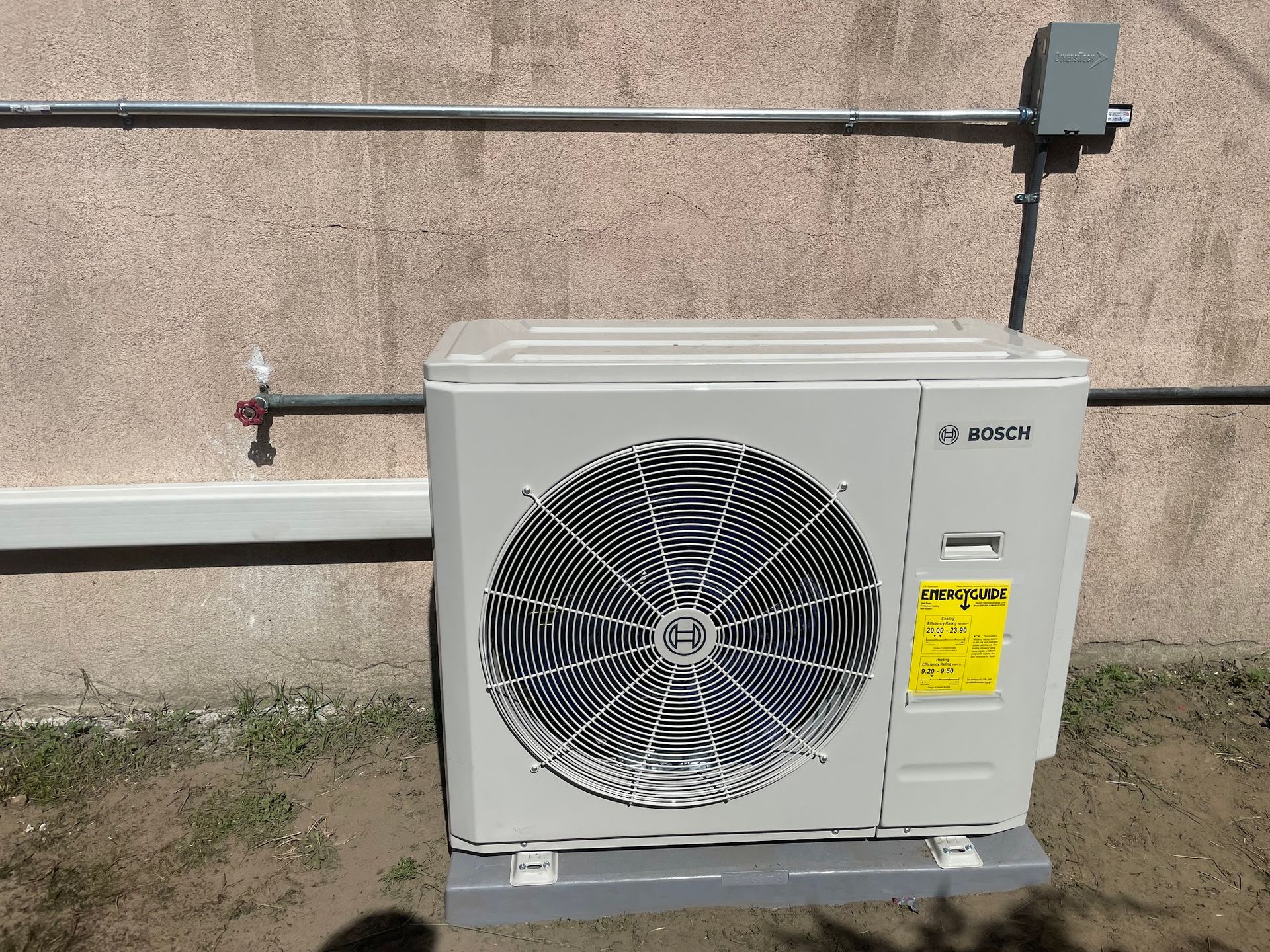 Outdoor air conditioning unit mounted on a concrete base, against a beige wall with electrical conduit.