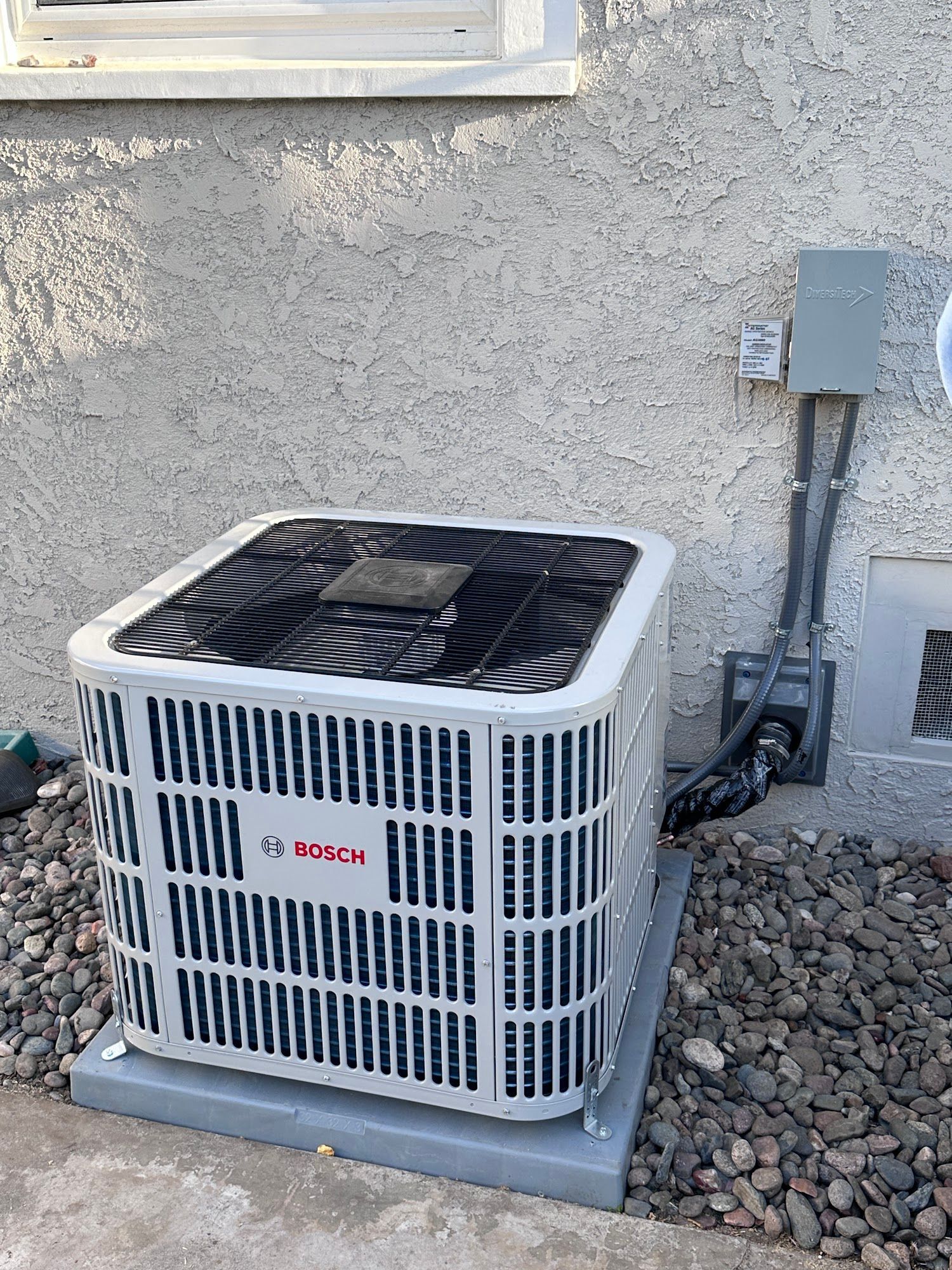 Bosch air conditioning unit, light grey and black, mounted on concrete pad next to a light-colored stucco wall.