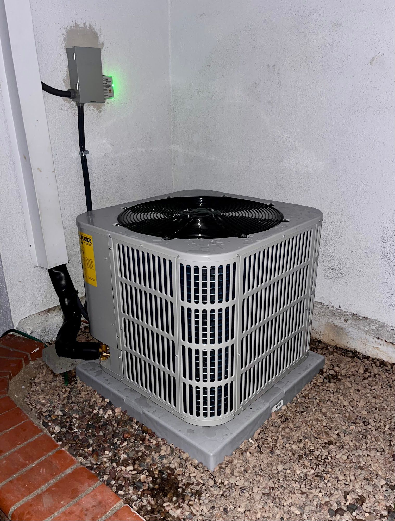 Air conditioning unit next to a white wall and brick foundation.