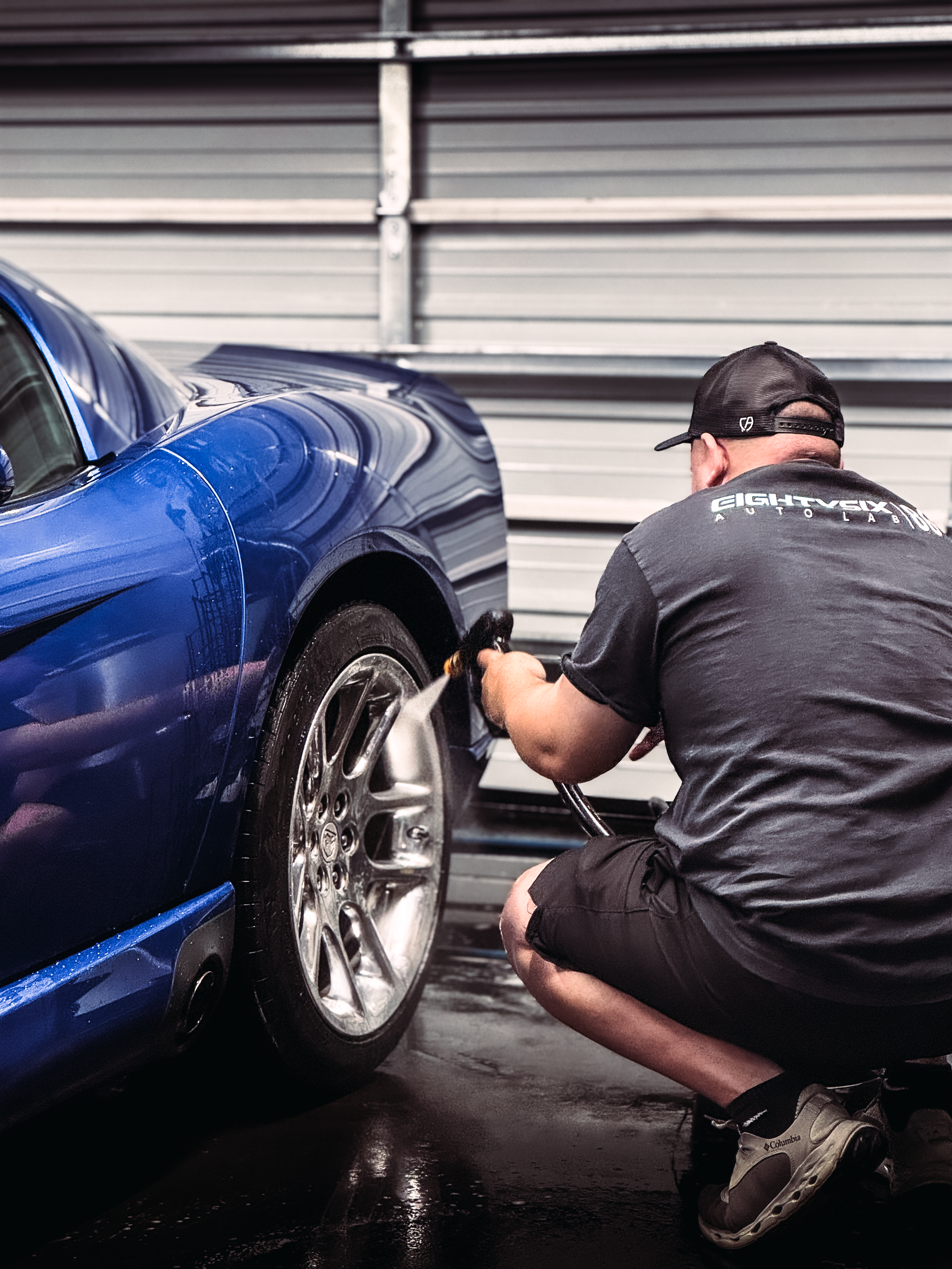 Man washes a blue sports car's tire with a spray hose near a garage door.