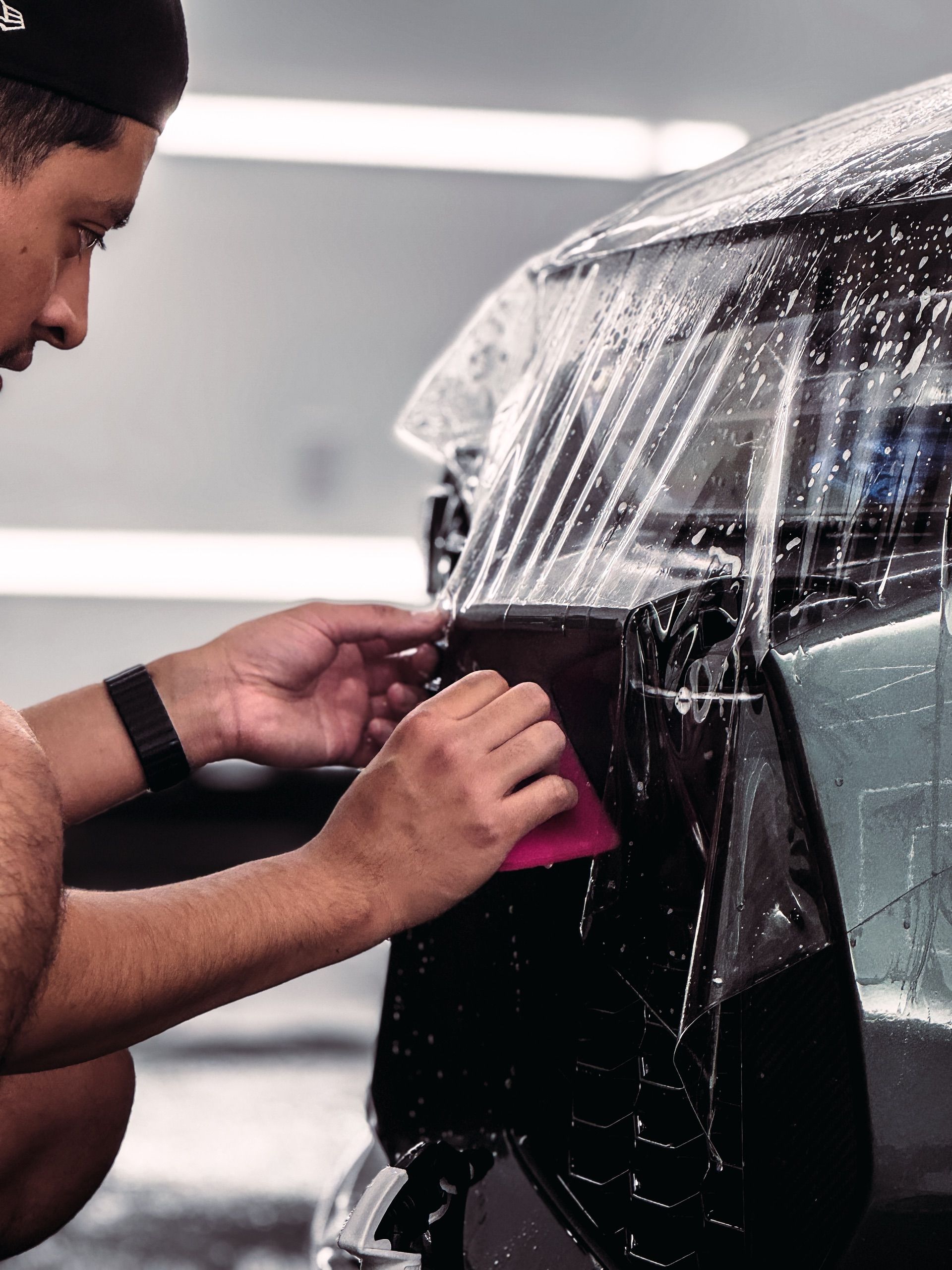 A person using a pink squeegee to smooth a clear protective film onto a wet car surface in a professional workshop.