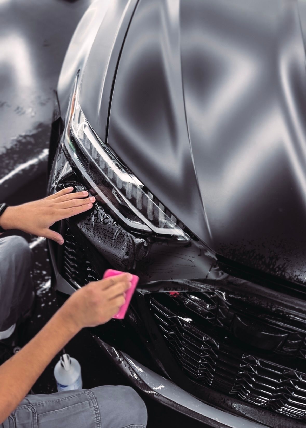 Hands applying protective film to a black car's grill, using a pink squeegee.