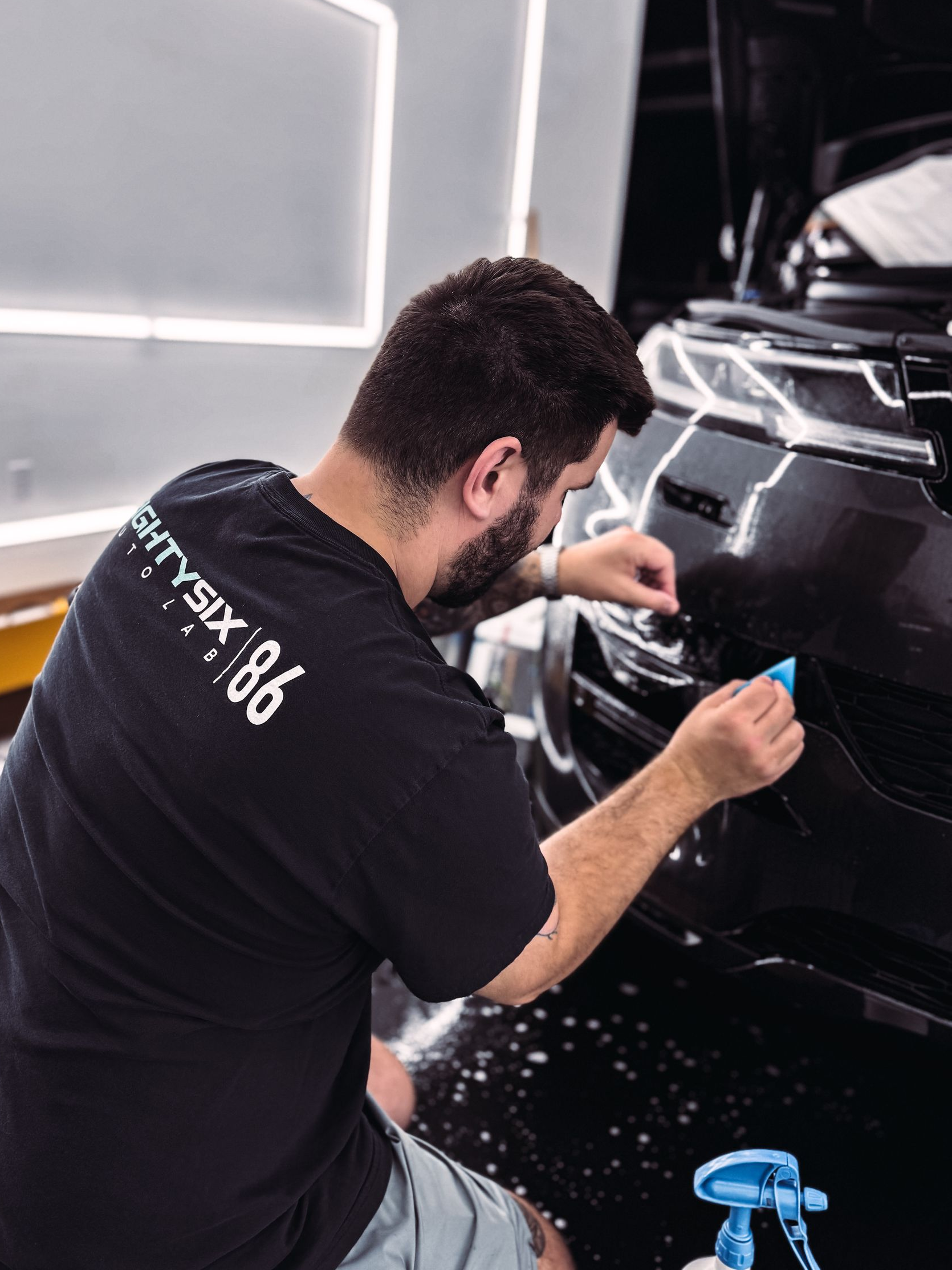 Man applying film to a car bumper in a workshop with bright lights.