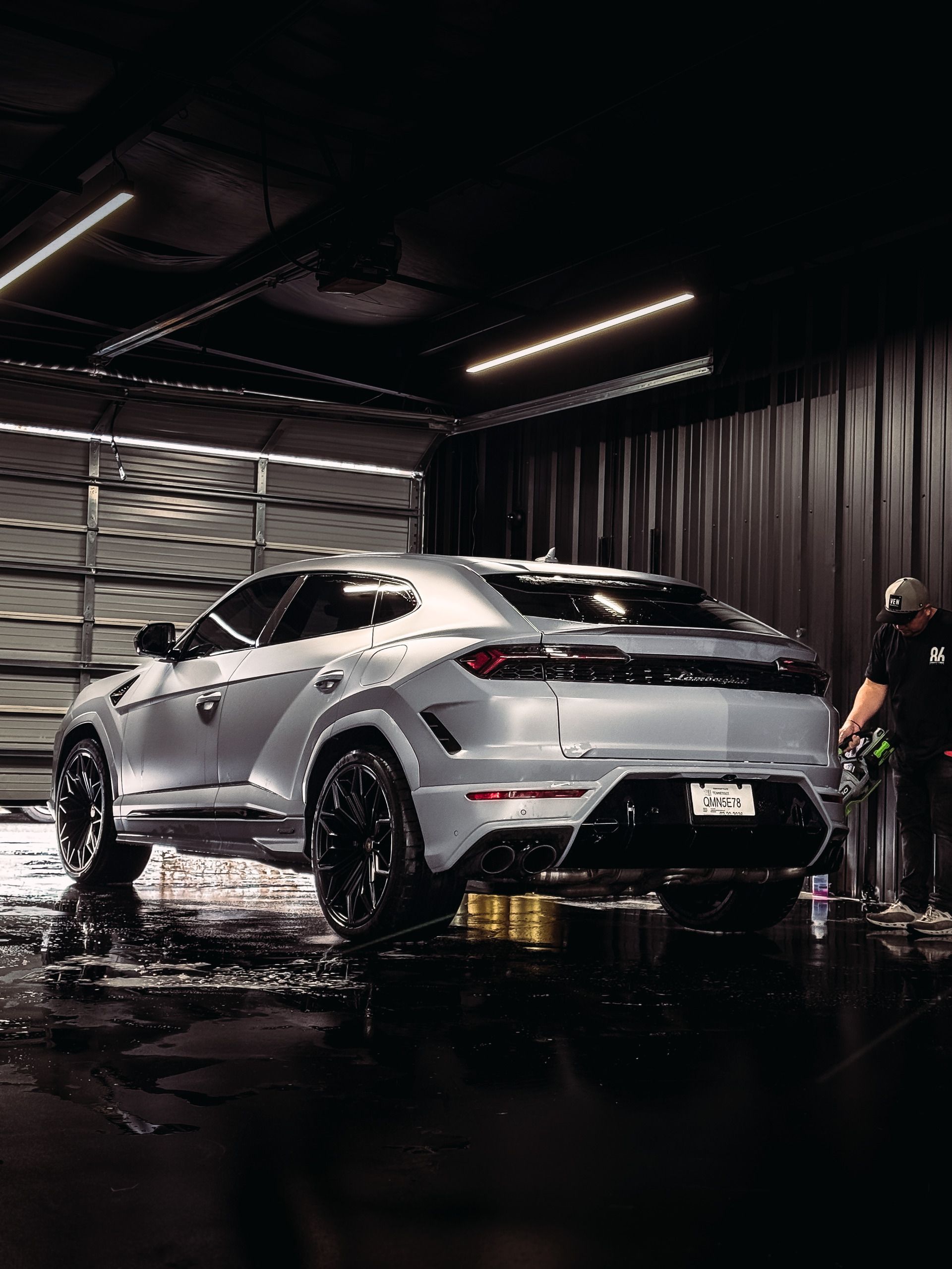 Silver Lamborghini SUV being washed by a person in a dark garage.