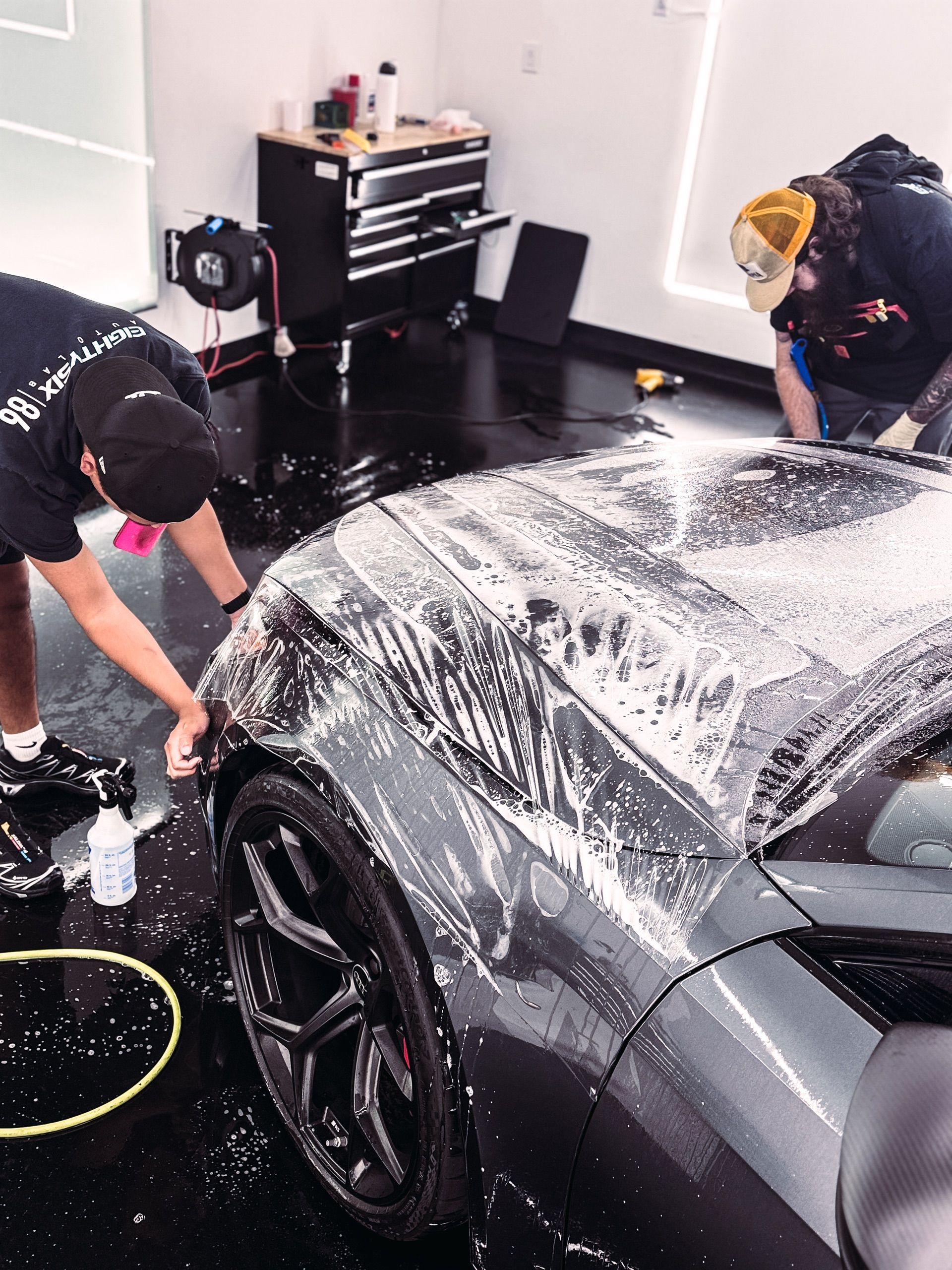 Two technicians wash a dark-colored car covered in white foam inside a well-lit workshop.