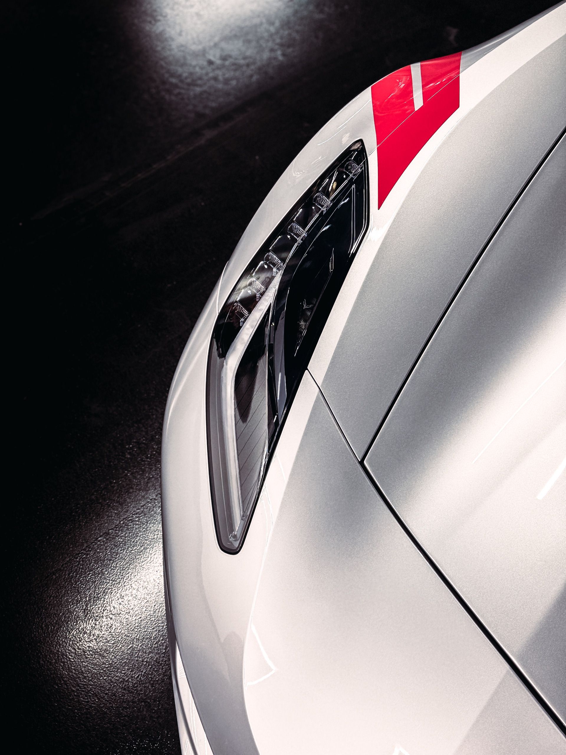 Close-up of a silver sports car headlight with a red stripe detail, angled against a dark background.
