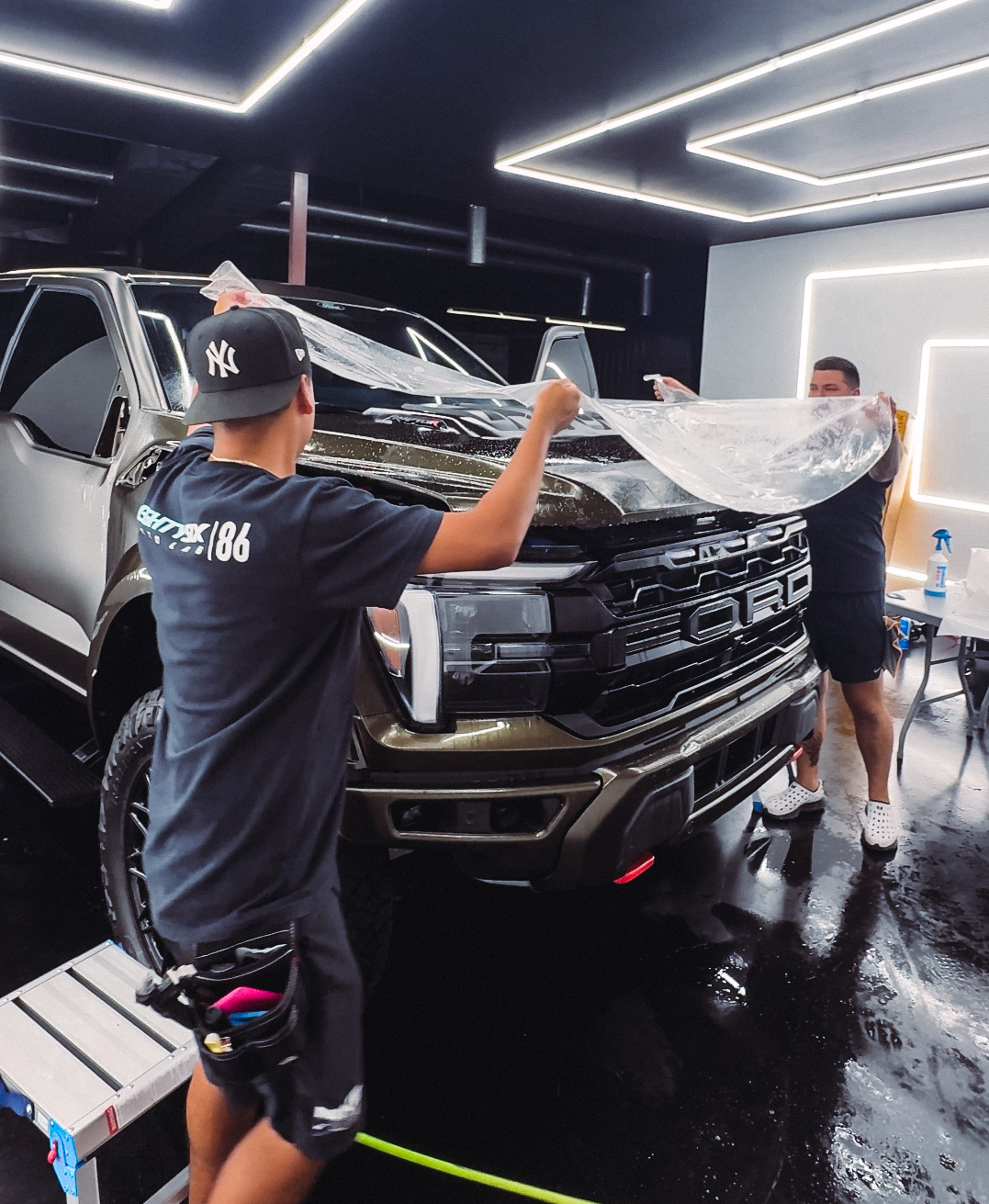 Two people applying protective film to the hood of a brown pickup truck inside a shop.