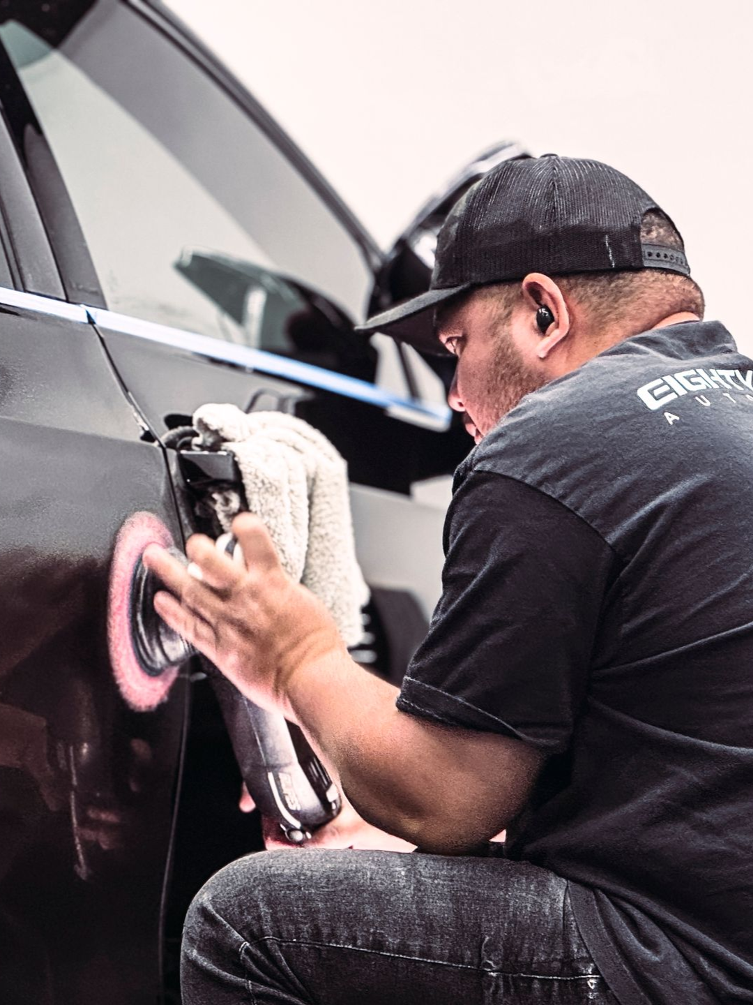 Man polishing a black car with a buffer, holding a towel.