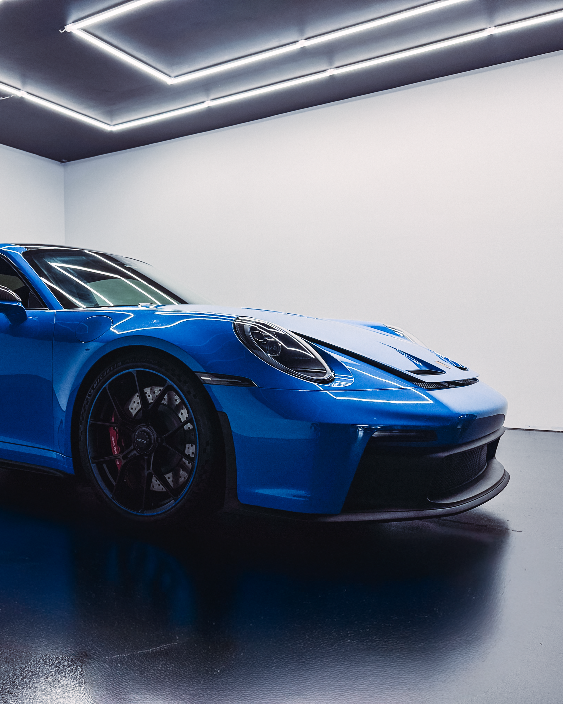 Blue Porsche sports car in a white-walled garage, with overhead neon lighting and black wheels.