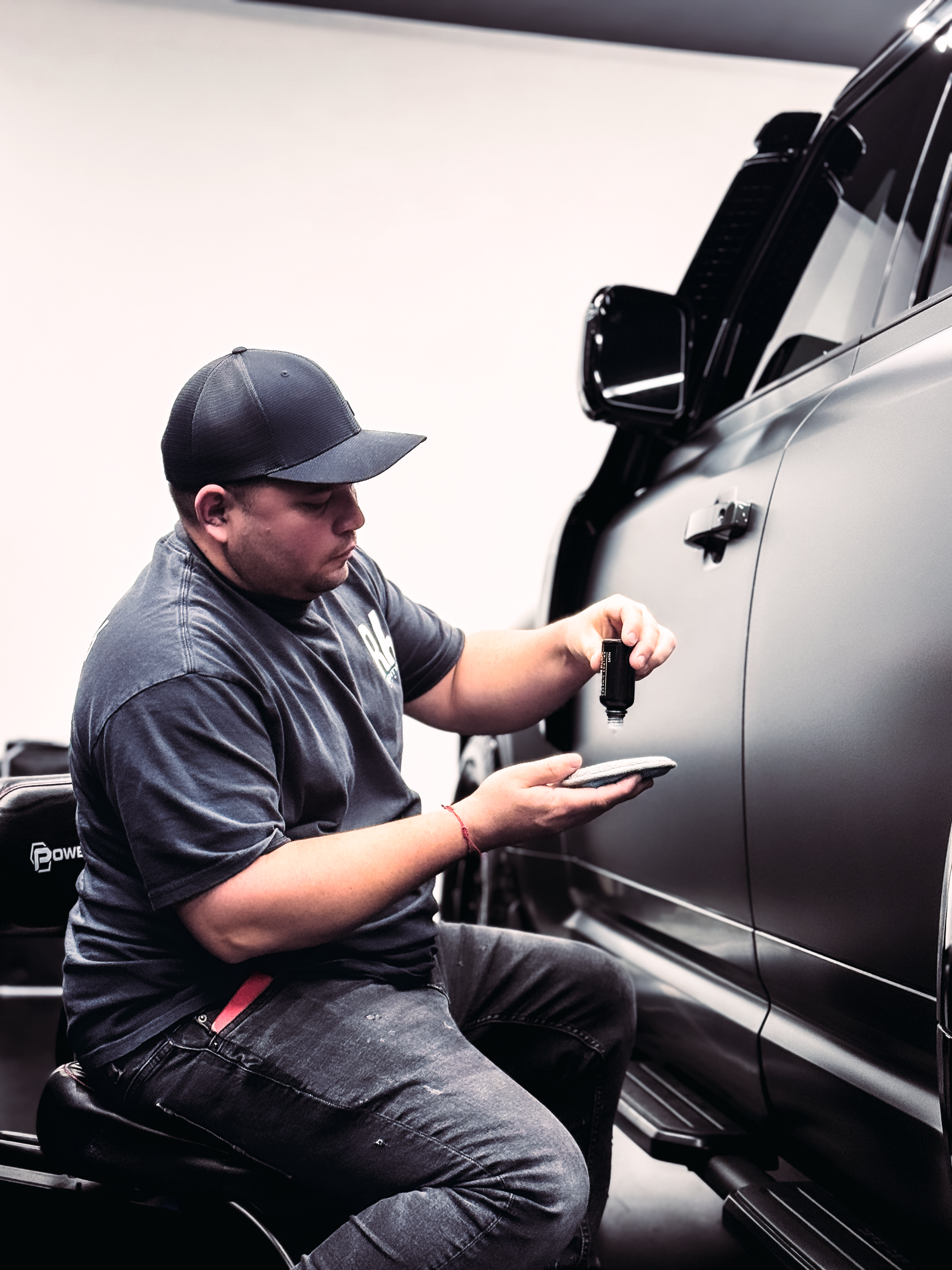 Man in a cap working on a matte black car door, holding a small part. Inside a shop.