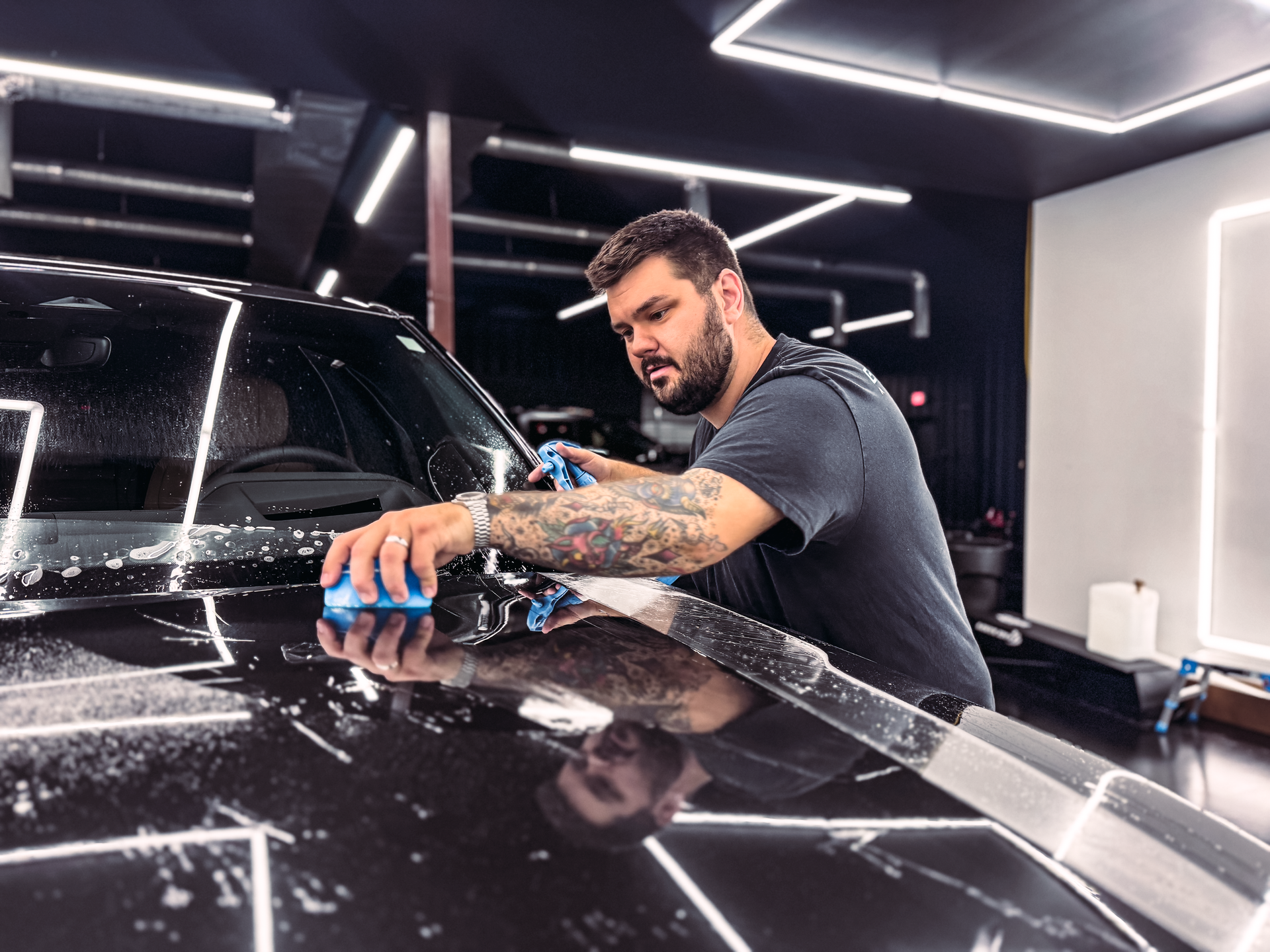 Man with tattoos washing a black car with a blue sponge in a well-lit garage.