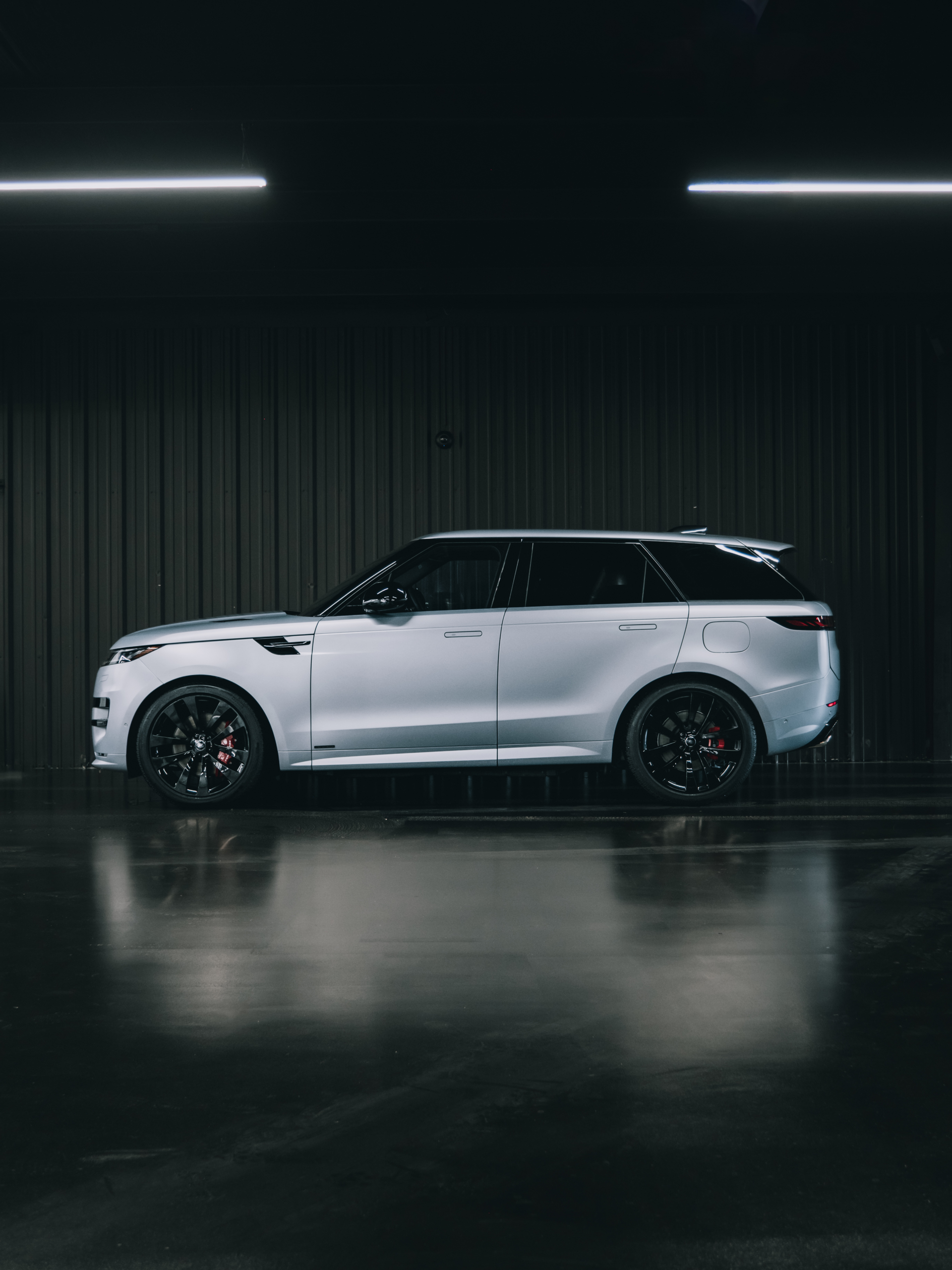 Silver Range Rover SUV parked indoors, black rims, under bright lights, dark background.