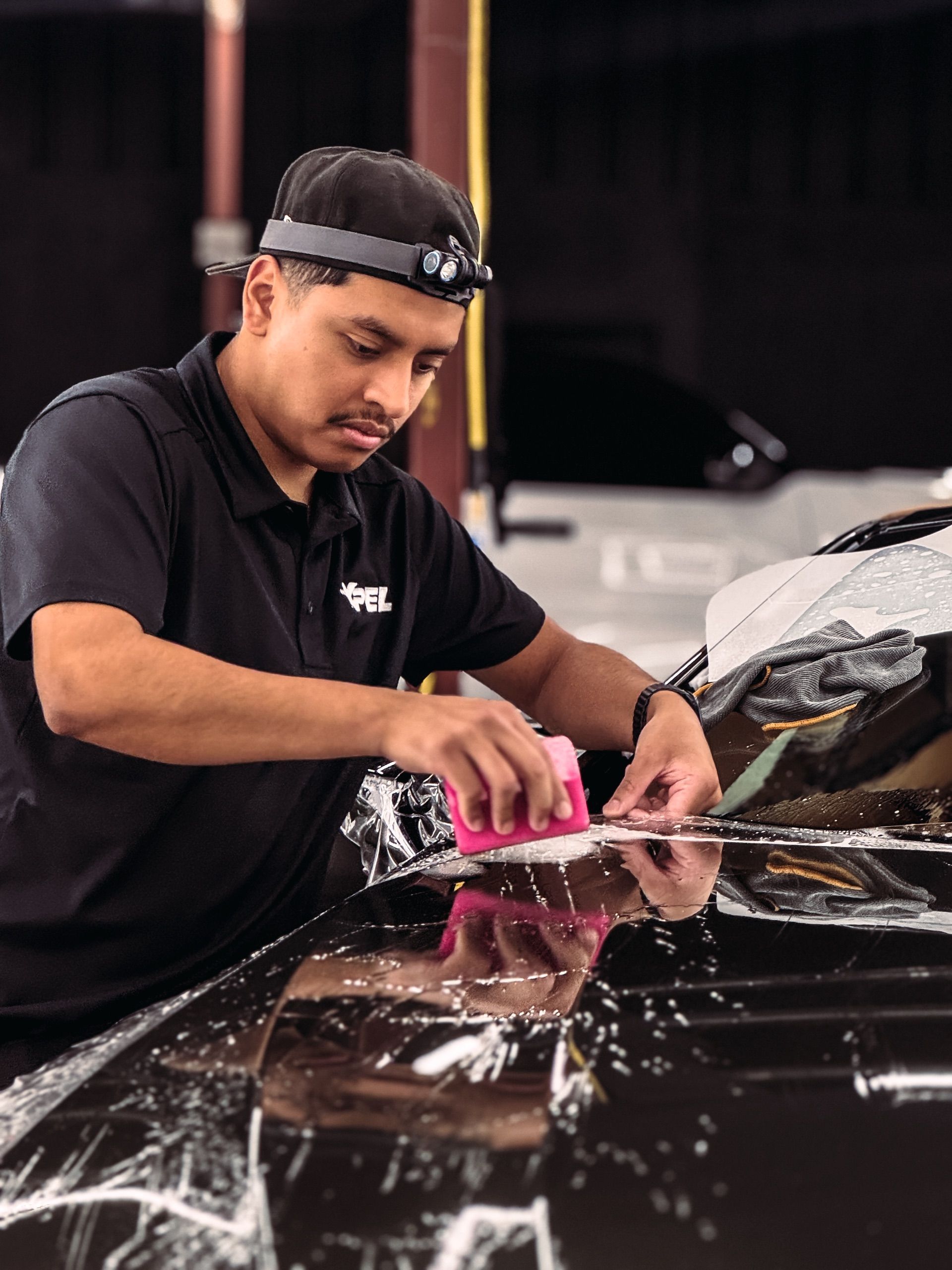 Person applying film to a car; indoors, using a squeegee, wearing a headlamp.