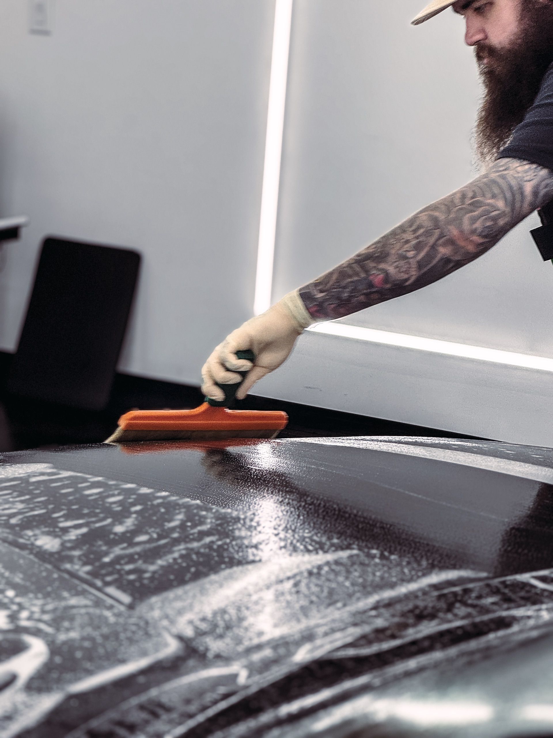 A person with a tattooed arm wears a glove and uses an orange squeegee to wipe soapy water off a dark car hood.