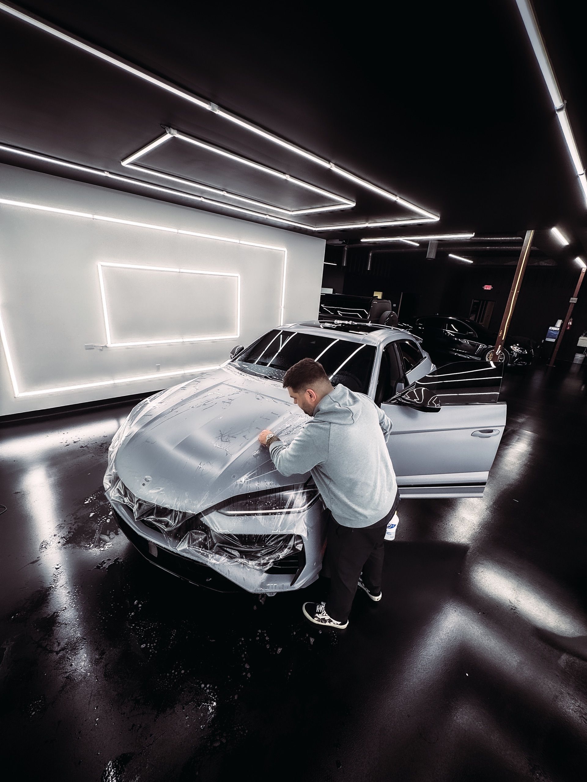 Man applying film to a silver car in a brightly lit garage.