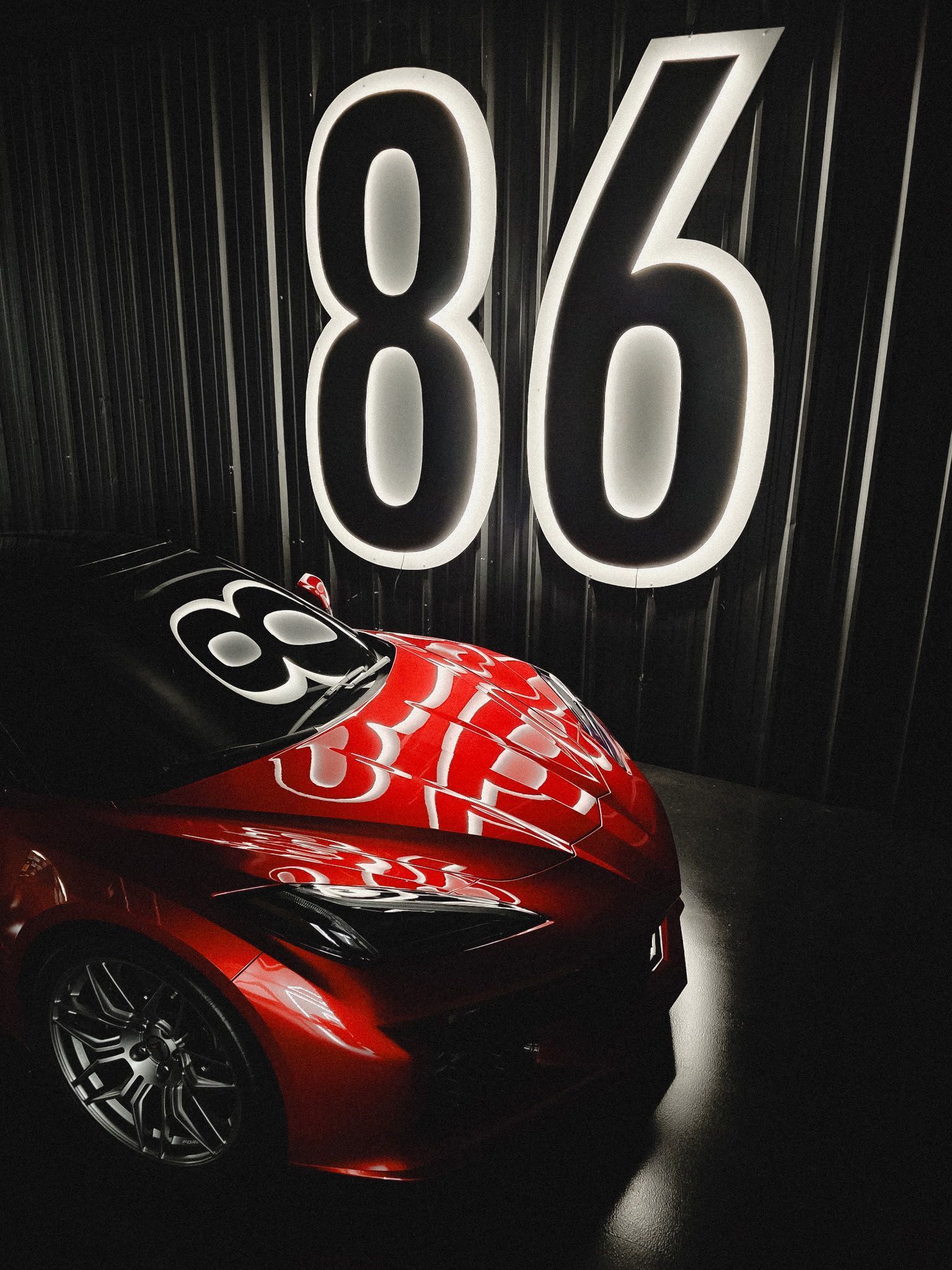 A glossy red sports car parked in a dark room beneath a large, glowing, illuminated number 86 on the wall.
