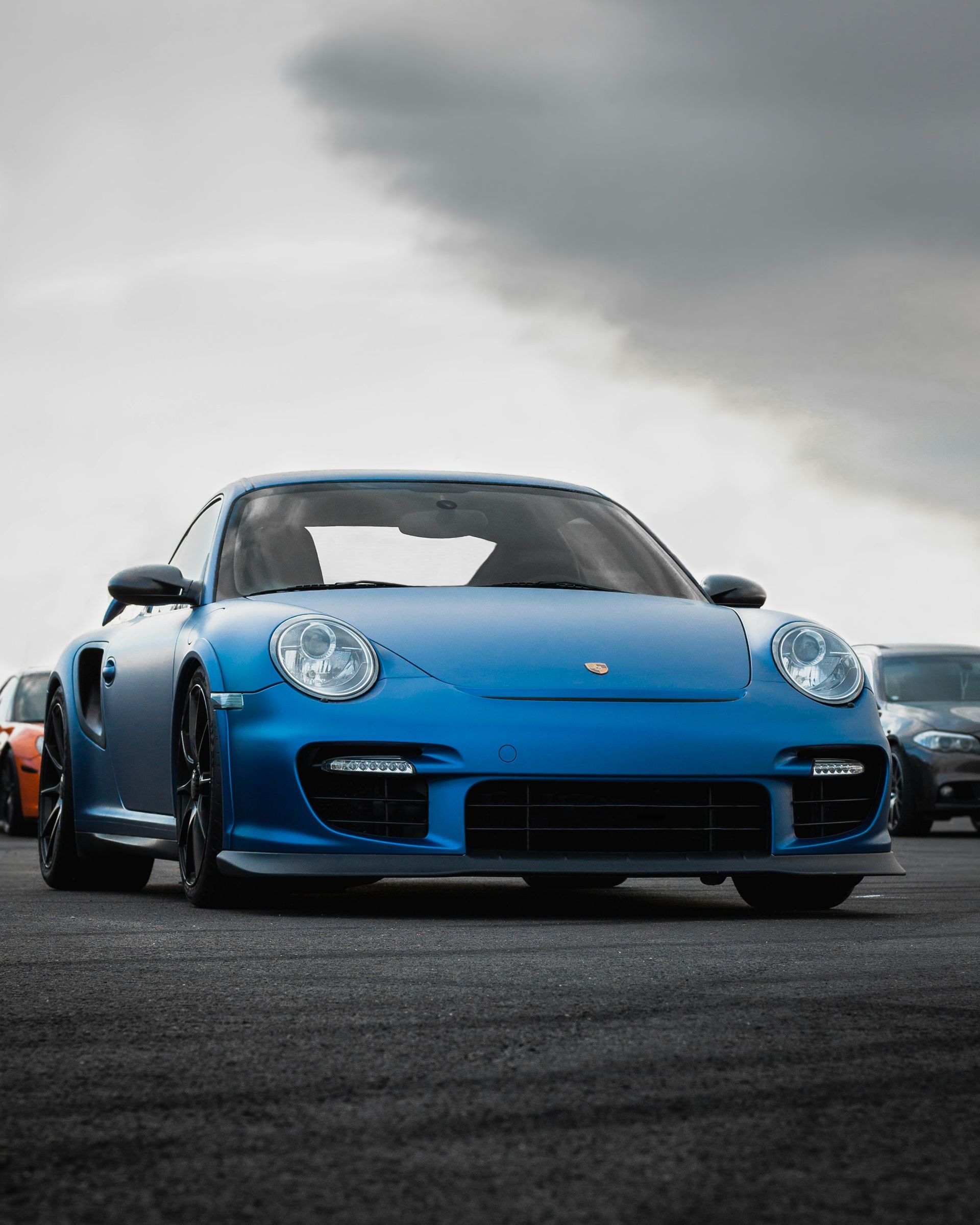 Blue Porsche sports car on asphalt under a cloudy sky, other cars in background.